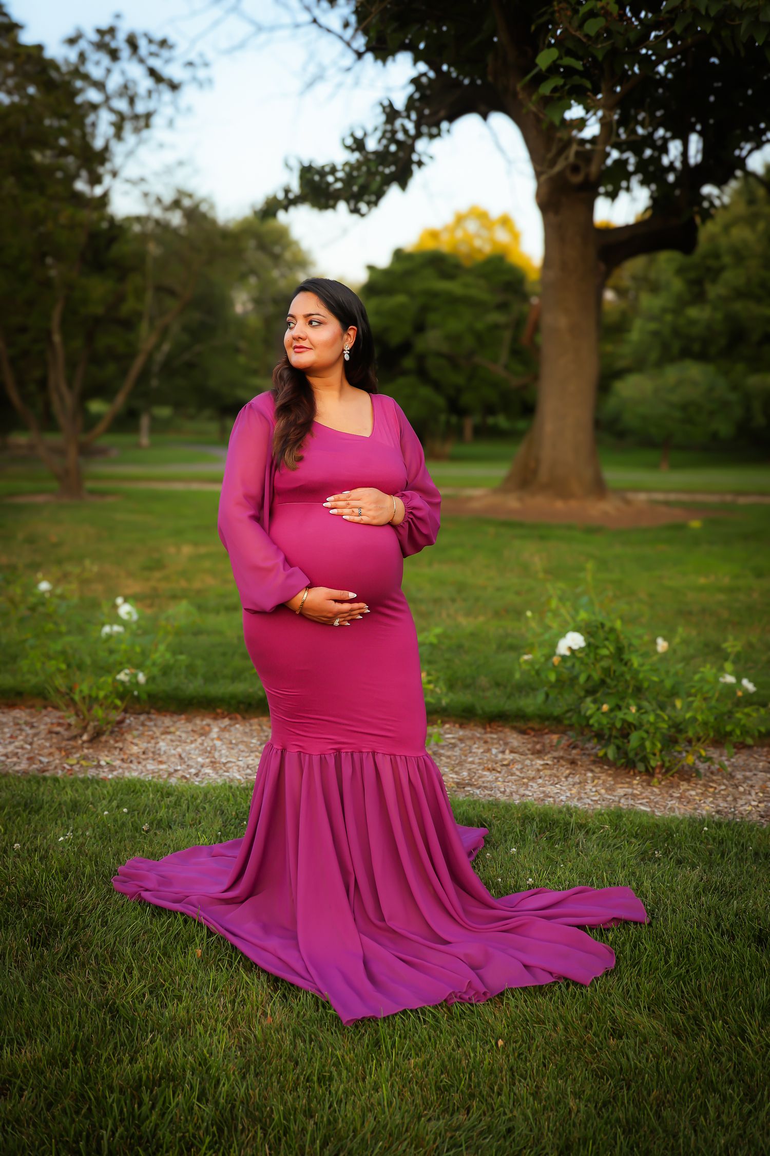 A person in a flowing magenta dress poses gracefully on grass in a park setting during golden hour.