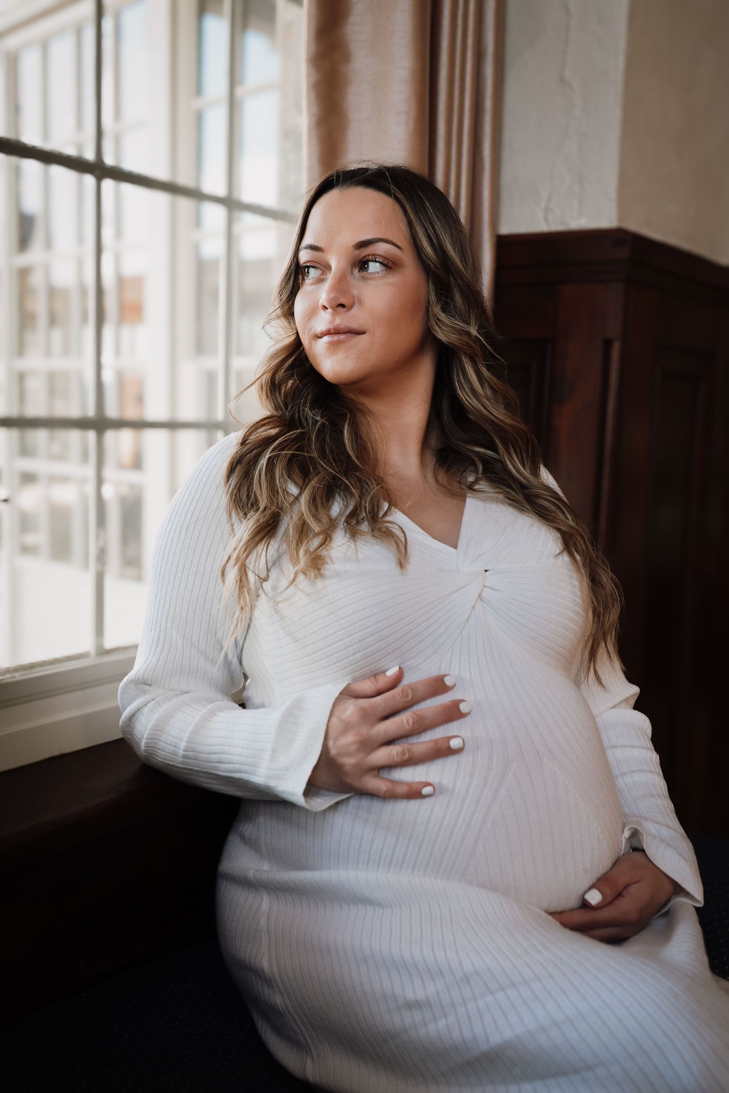 A pregnant woman in a white knit sweater sits by a window in natural light during a maternity photoshoot.