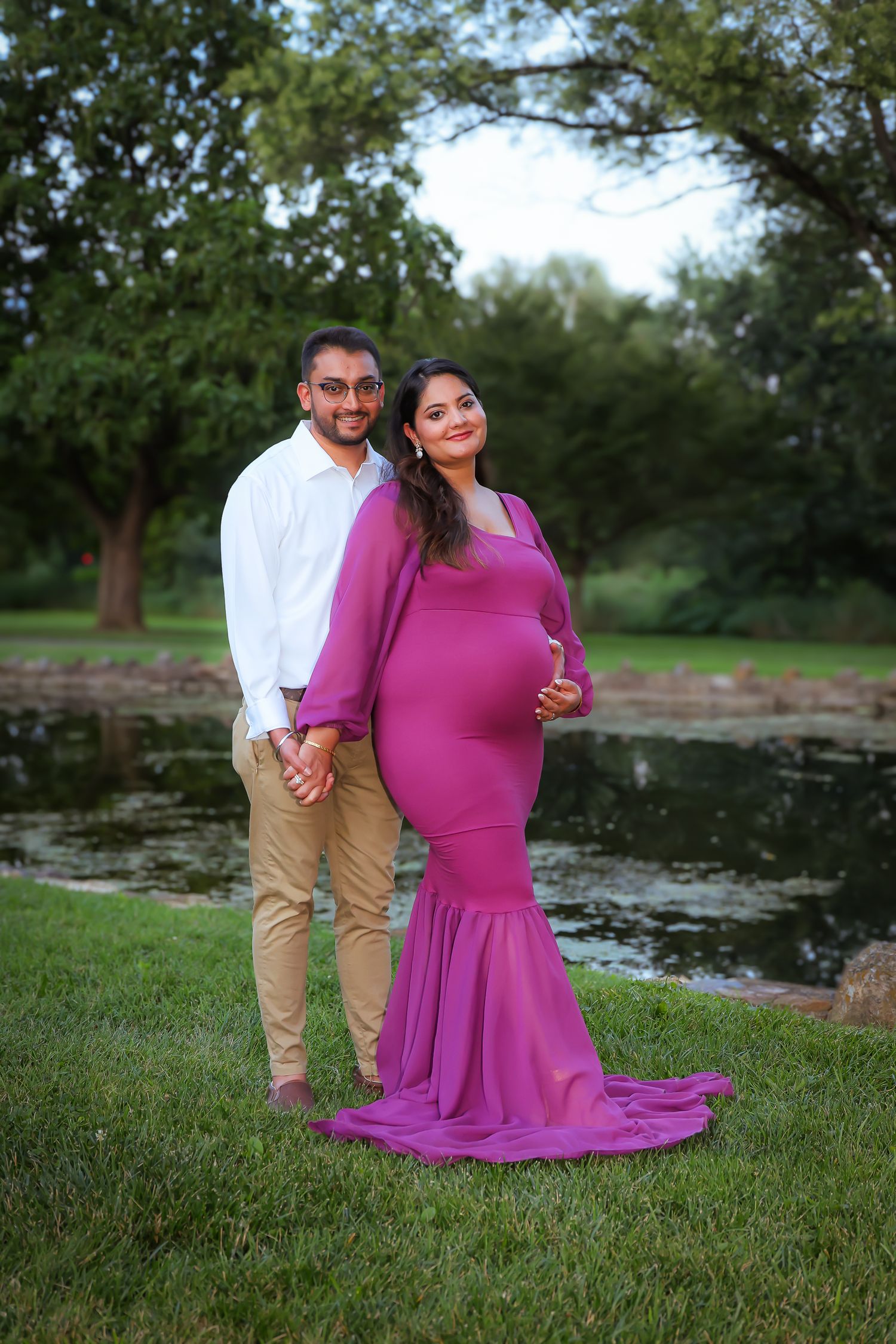 A couple stands together near a pond, with the person in a flowing magenta dress creating an elegant silhouette at sunset.