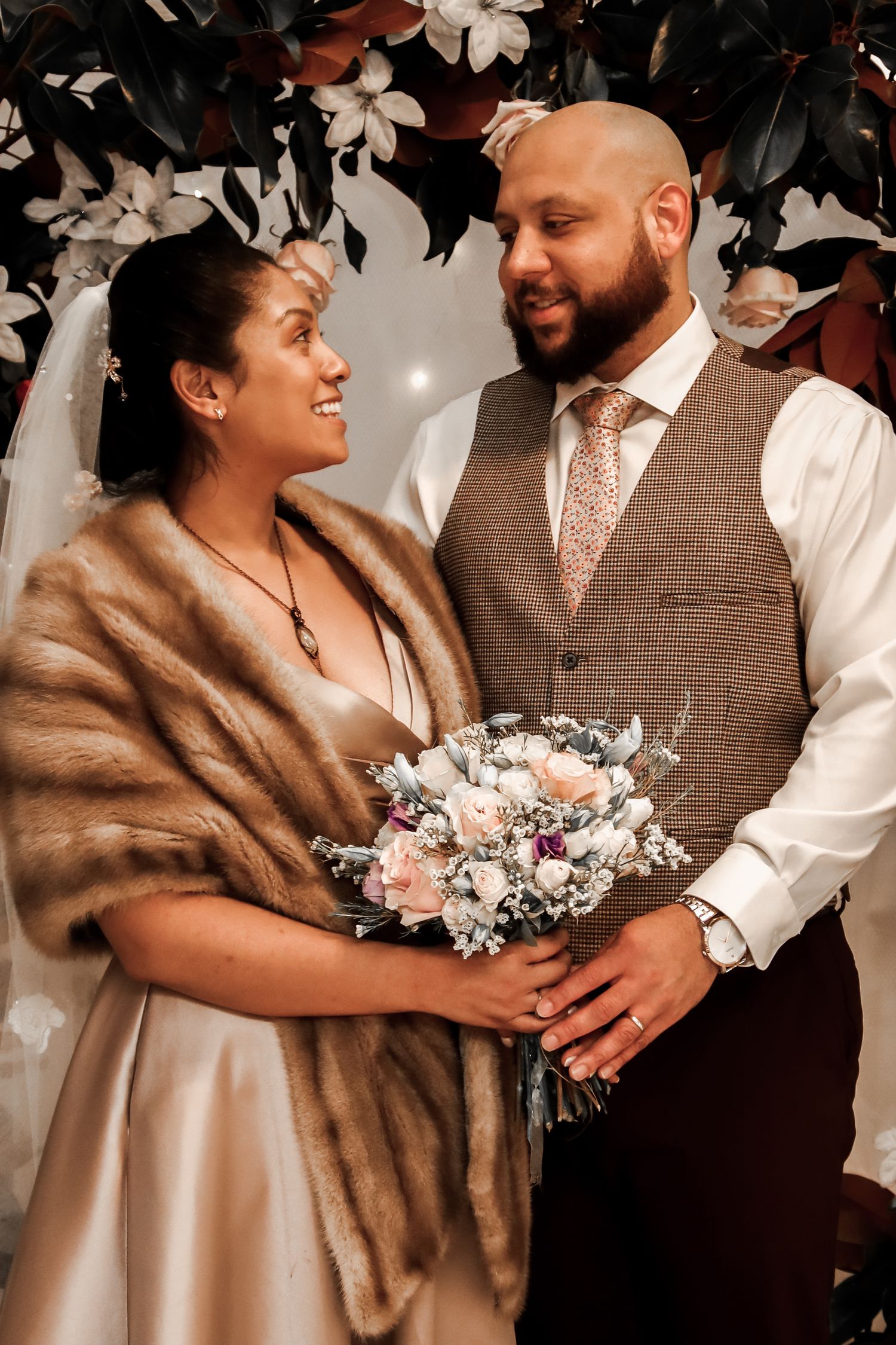 A couple shares intimate moments in wedding attire with a fur wrap and floral bouquet in a romantic indoor setting.