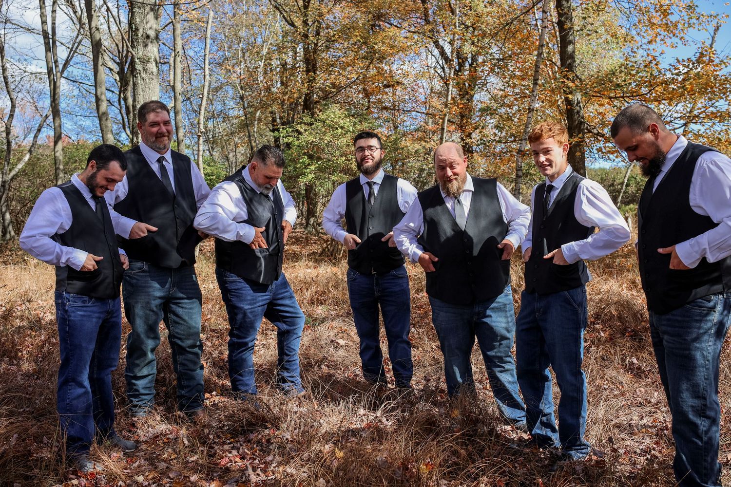 Groomsmen in navy suits pose playfully in a fall forest setting during a wedding photoshoot.