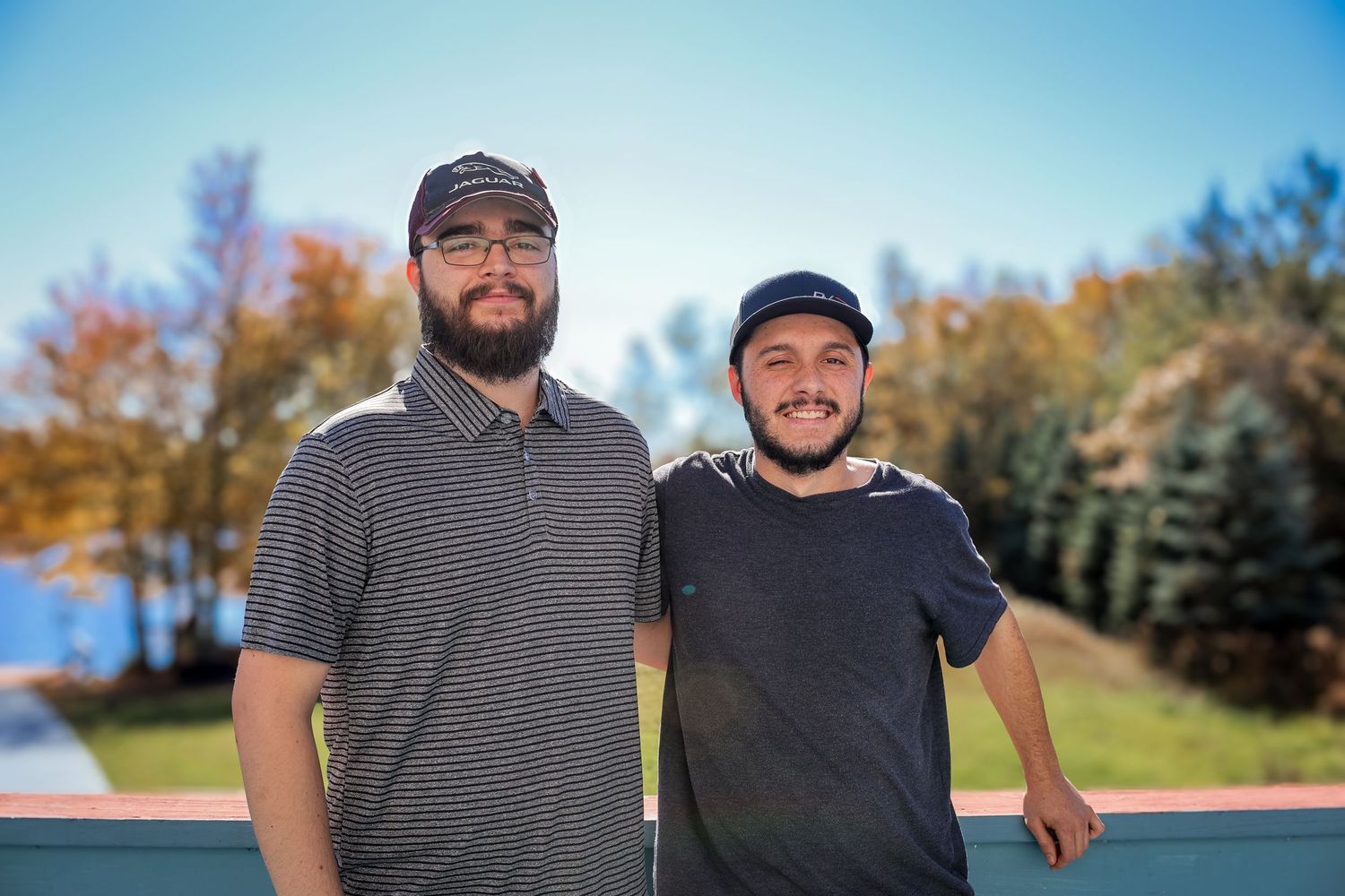 Two golfers stand together on a sunny golf course with autumn trees in the background.