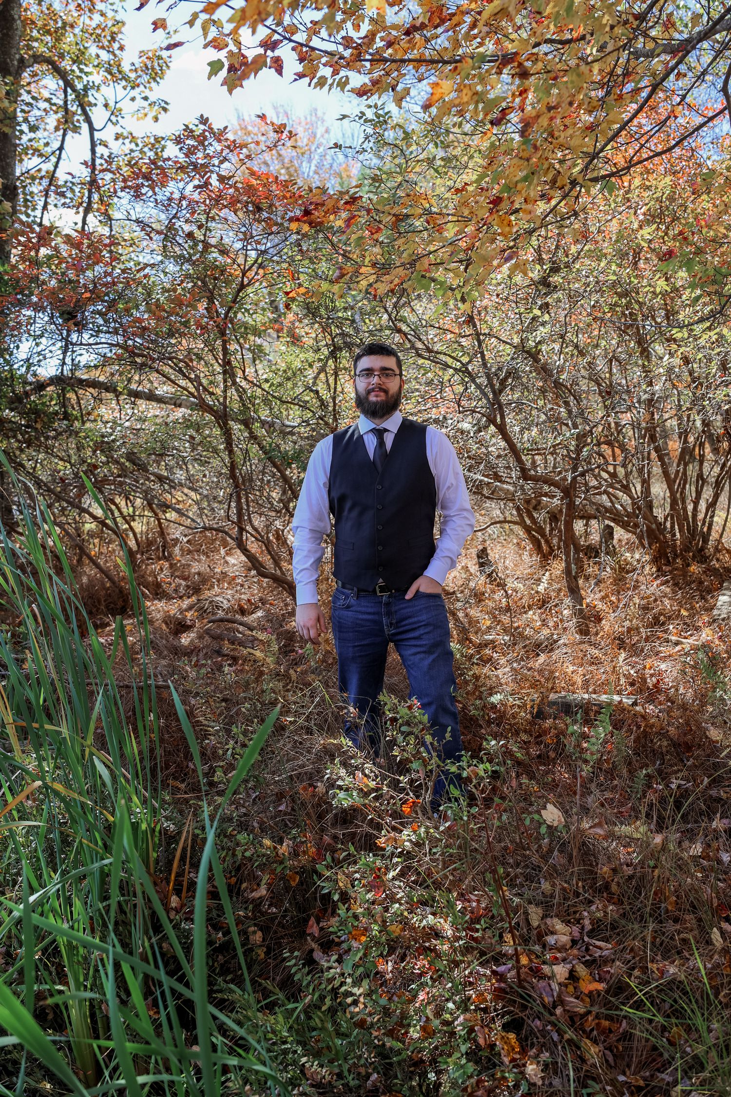 Someone wearing a navy vest and white shirt stands among autumn foliage and tall grass on a wooded hillside.
