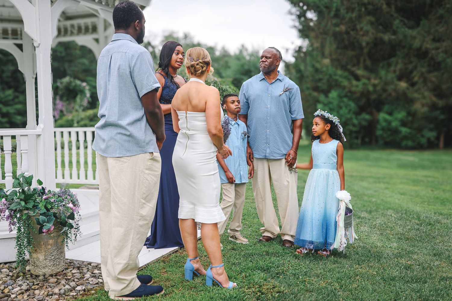 Wedding guests mingle on a lush green lawn outside a white gazebo during a summer celebration.