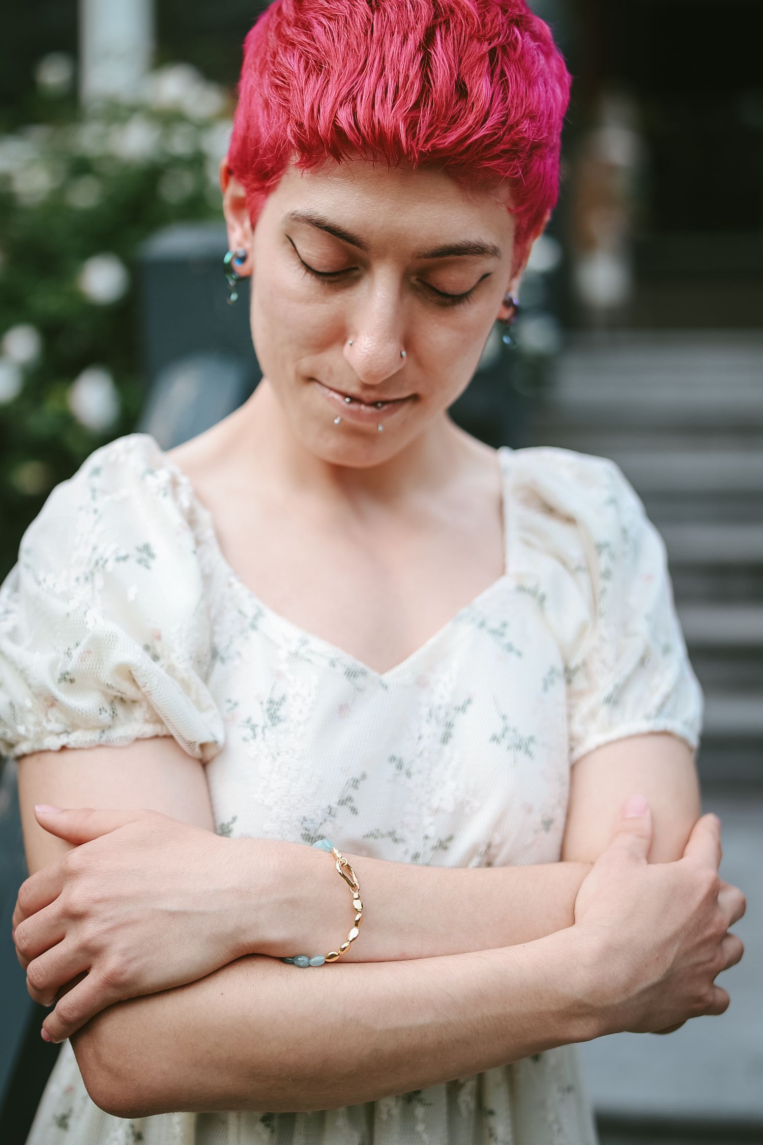 A person with vibrant pink short hair wearing a delicate floral dress stands with arms crossed in a contemplative pose.