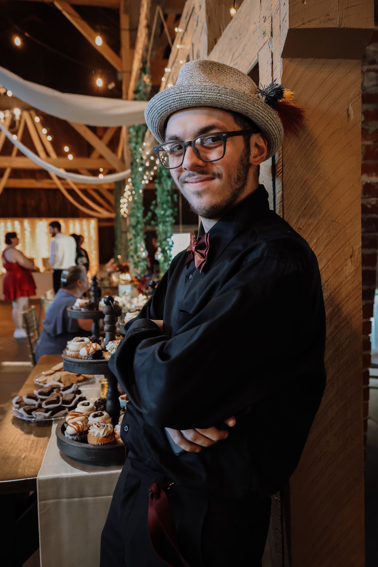 A stylishly dressed person in a hat and glasses stands by a dessert table at an event with twinkling lights and decor.