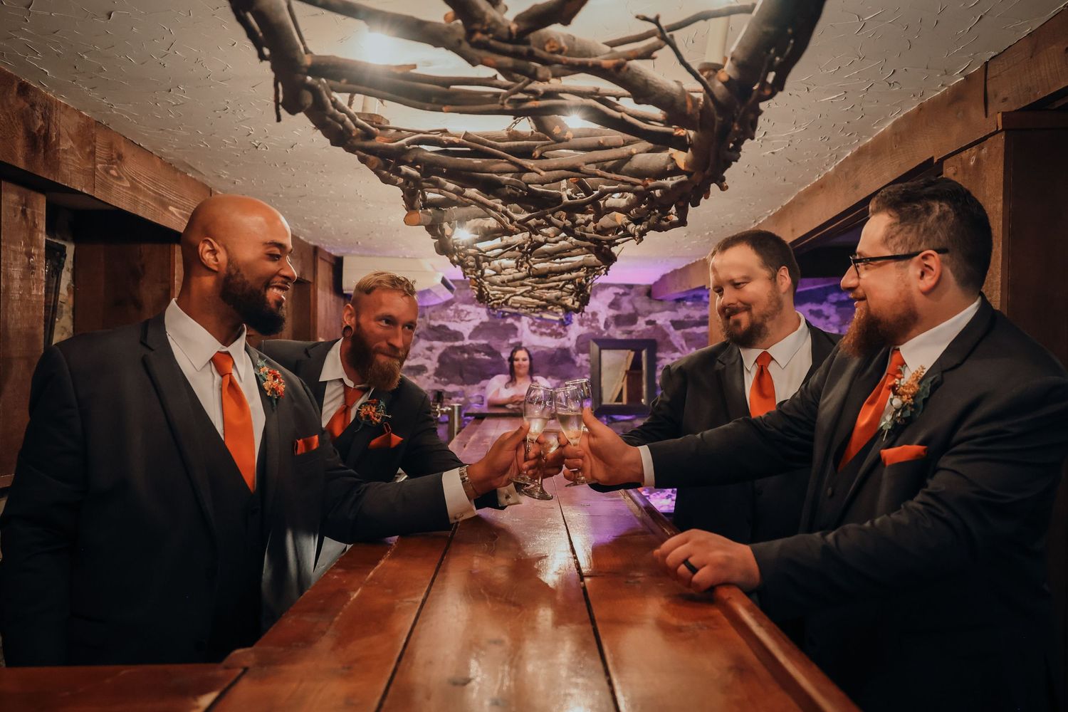 Groomsmen in matching black suits and orange ties toast with champagne glasses at a rustic wooden bar.