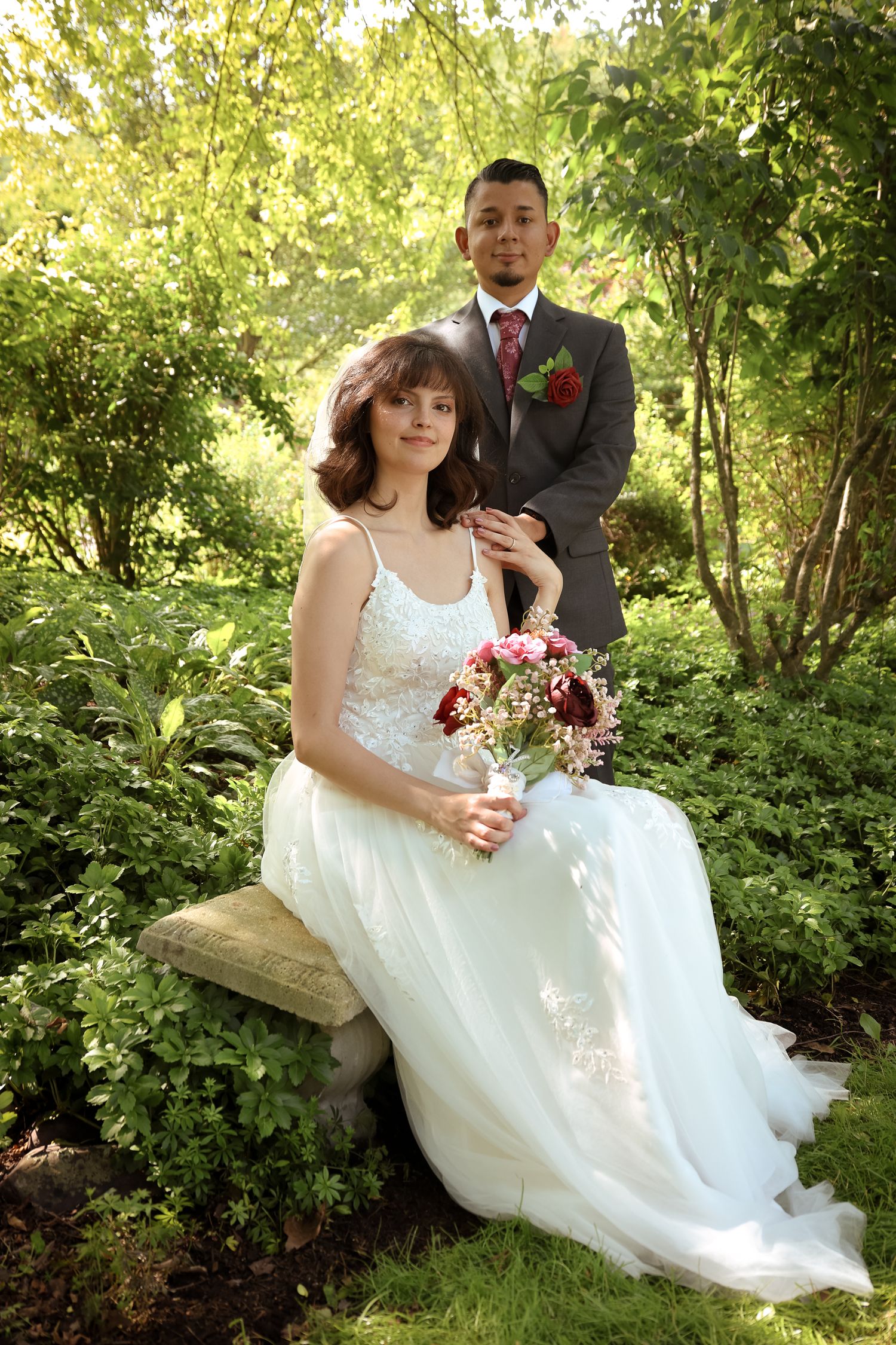A couple in wedding attire poses together on a rustic wooden bench surrounded by lush green foliage in a garden setting.