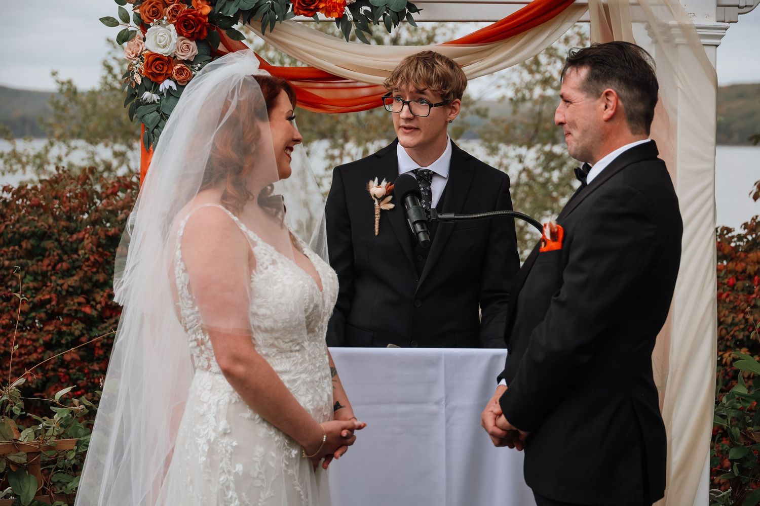 A couple exchanges vows during an outdoor wedding ceremony beneath a white arch decorated with red flowers.
