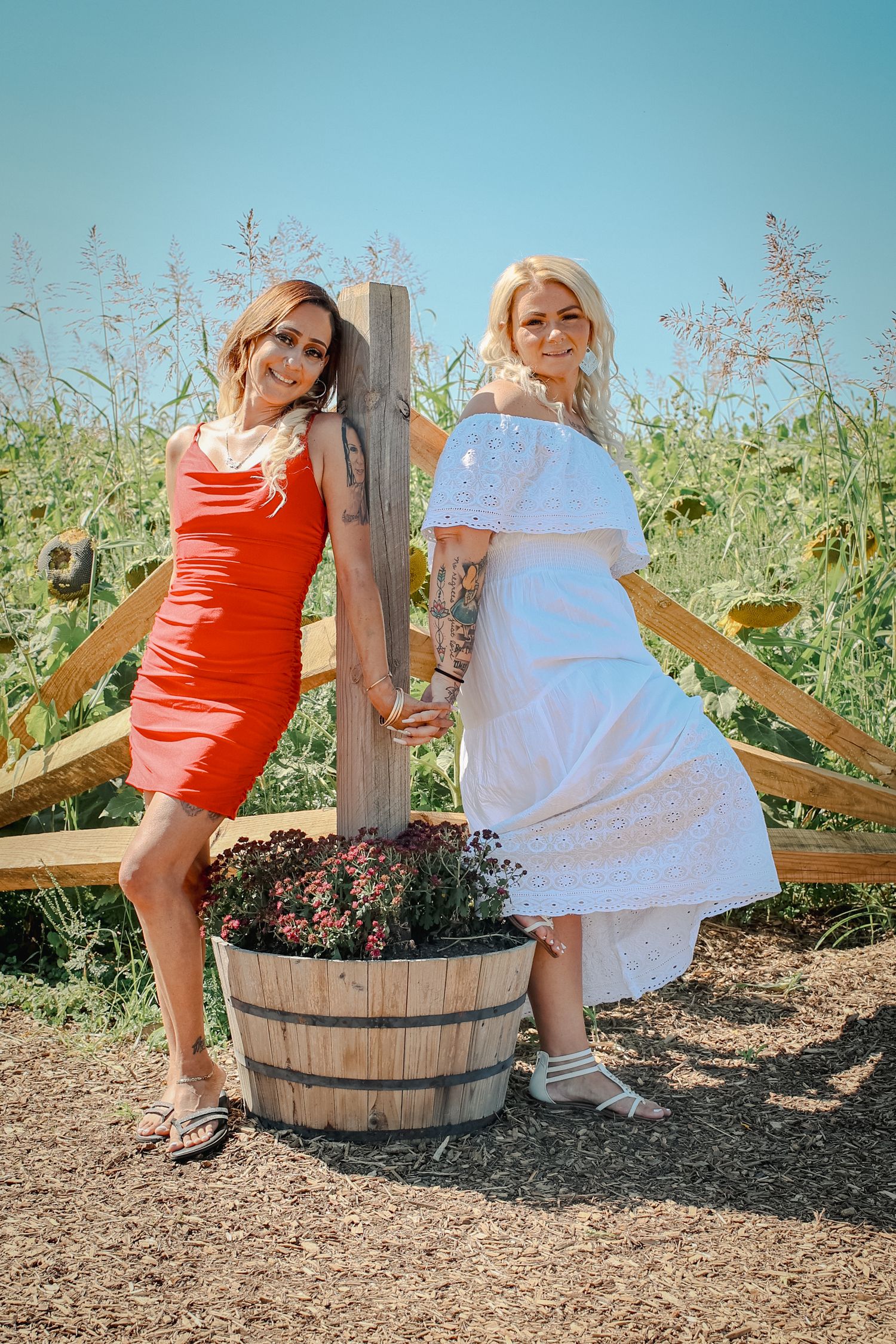 Two women pose by wooden fence, one in red dress and one in white summer dress on sunny day.