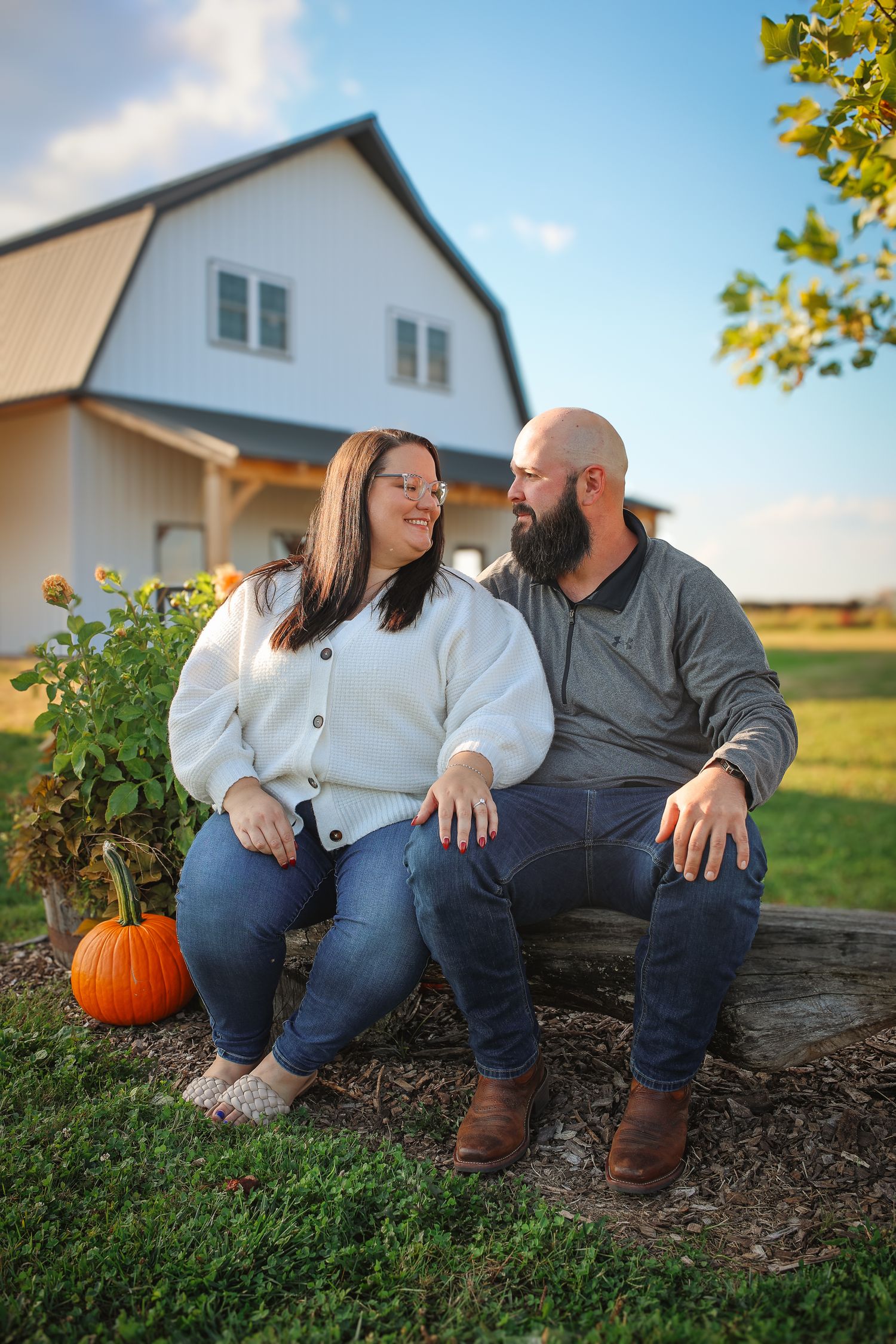 A couple sits together on a rustic log bench outside a barn with a pumpkin in autumn.