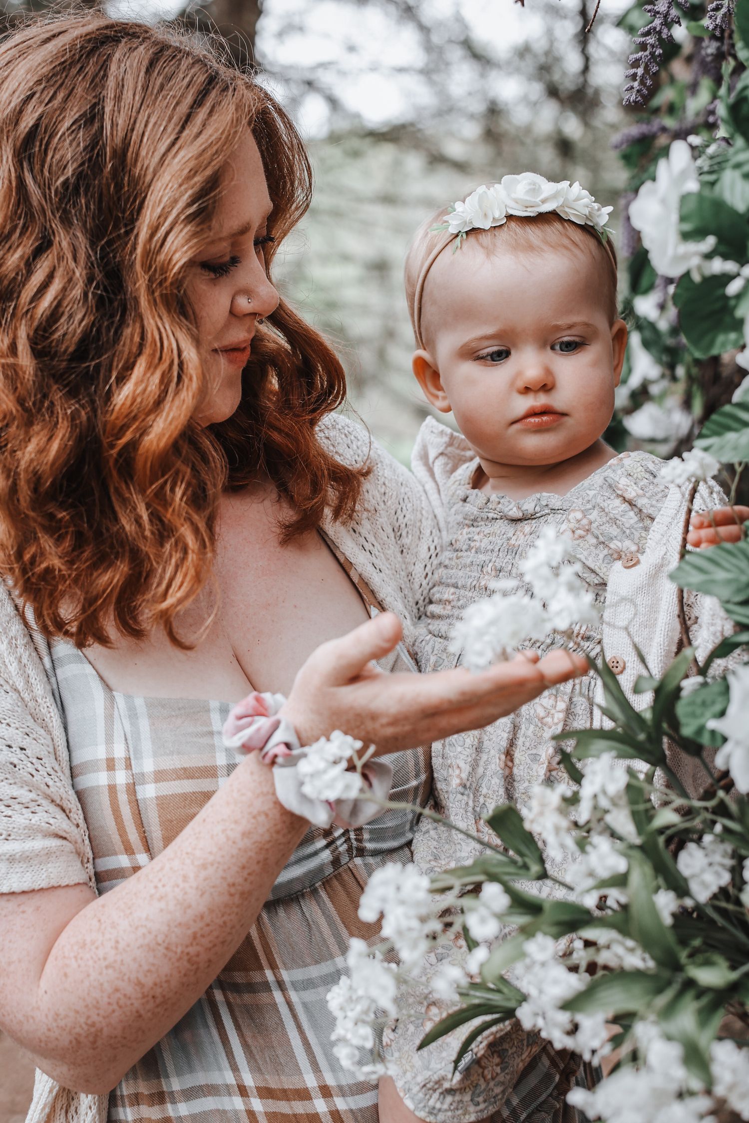 A mother and baby in matching neutral tones pose together amid blooming white spring flowers in a dreamy outdoor setting.