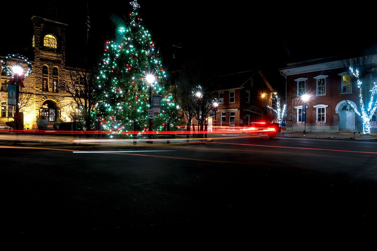 Illuminated Christmas tree and buildings with light trails from passing cars in a nighttime town square scene.