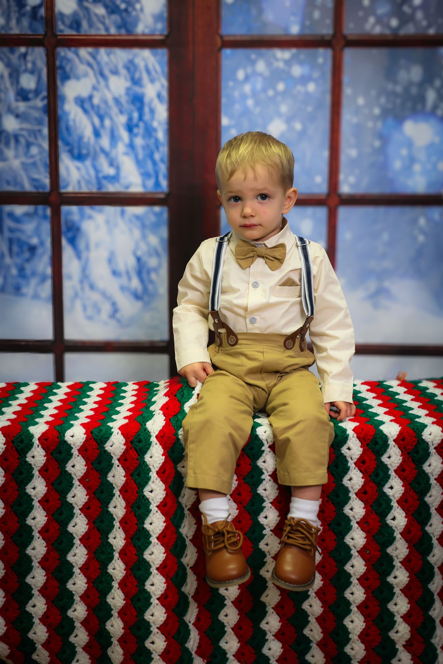 Small child sits on a festive red and green chevron blanket against a window backdrop with snowy winter scenery.