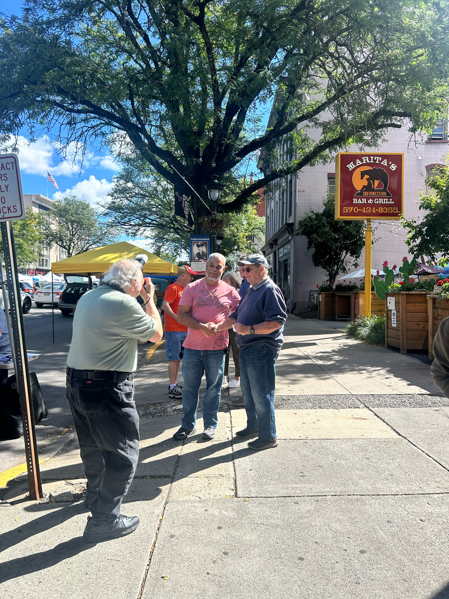 People gathered outside a small storefront with trees casting shade on a sunny afternoon.