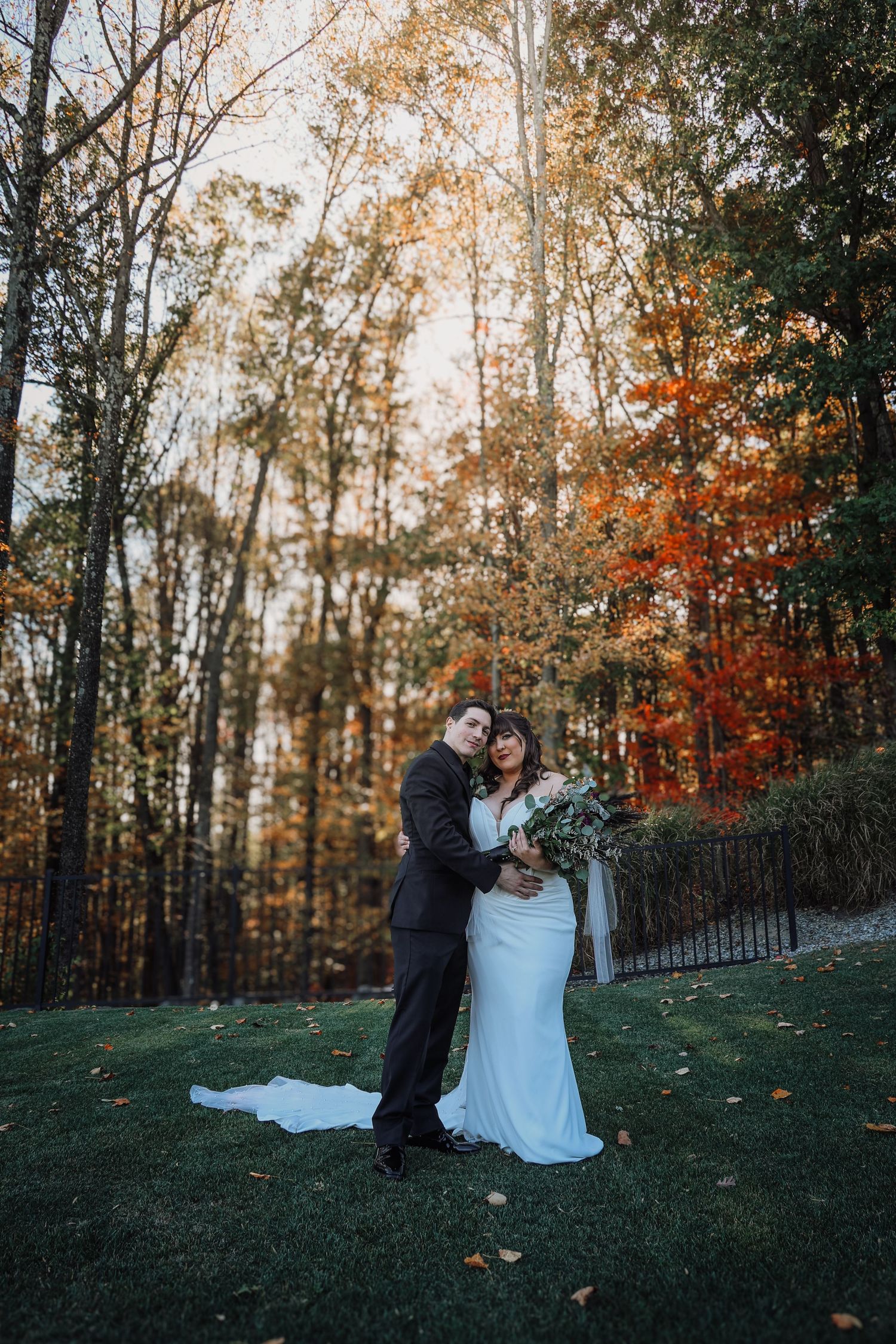 Couple in formal attire embraces on a grassy hillside during autumn with colorful fall foliage in the background.