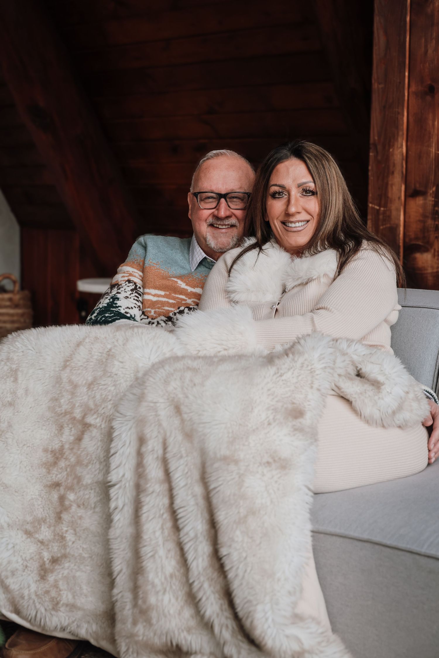 A couple snuggled together on a couch sharing a cozy white blanket while smiling and laughing.