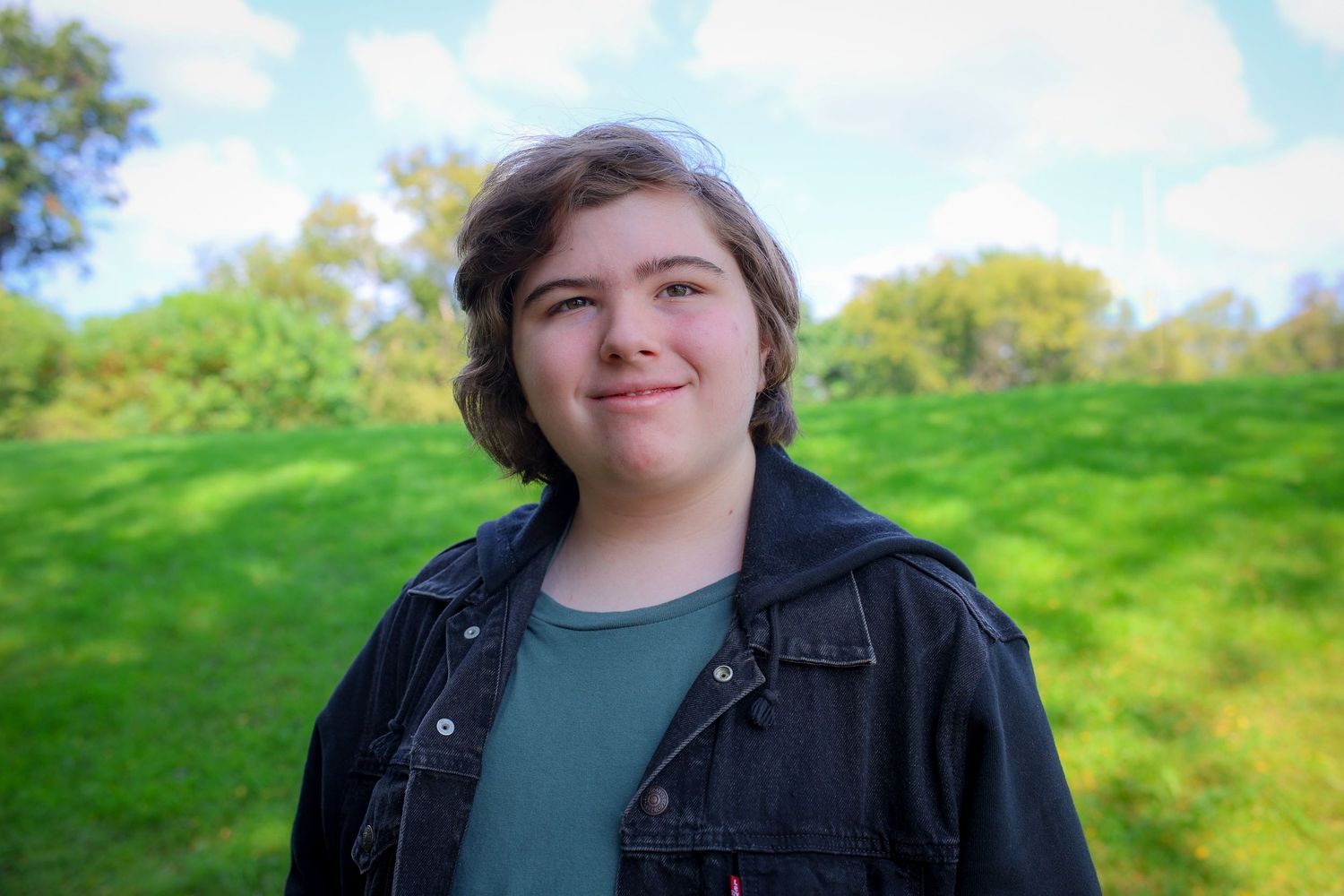 Someone wearing a black denim jacket smiles while standing in a grassy field on a sunny day.