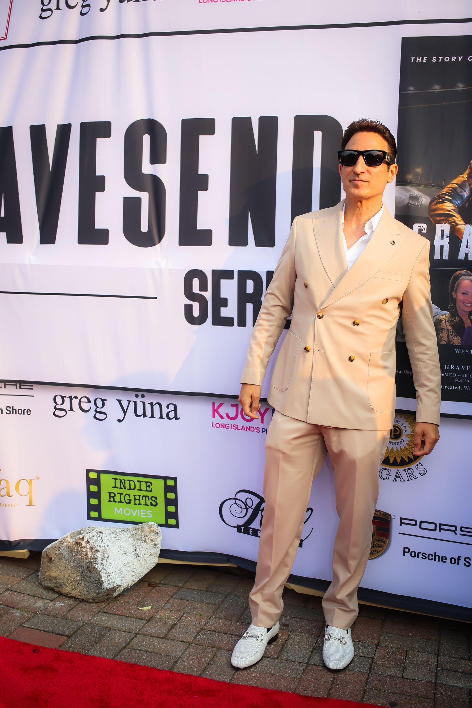 A person in a cream-colored suit and white sneakers poses against a backdrop with sponsor logos and text.