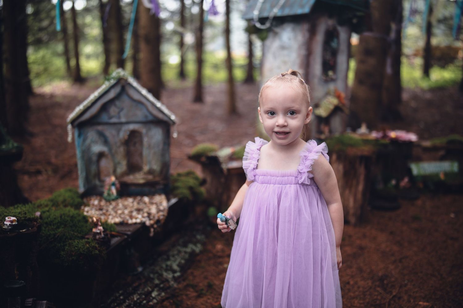 A child in a lavender dress stands among tall trees in a wooded forest setting.