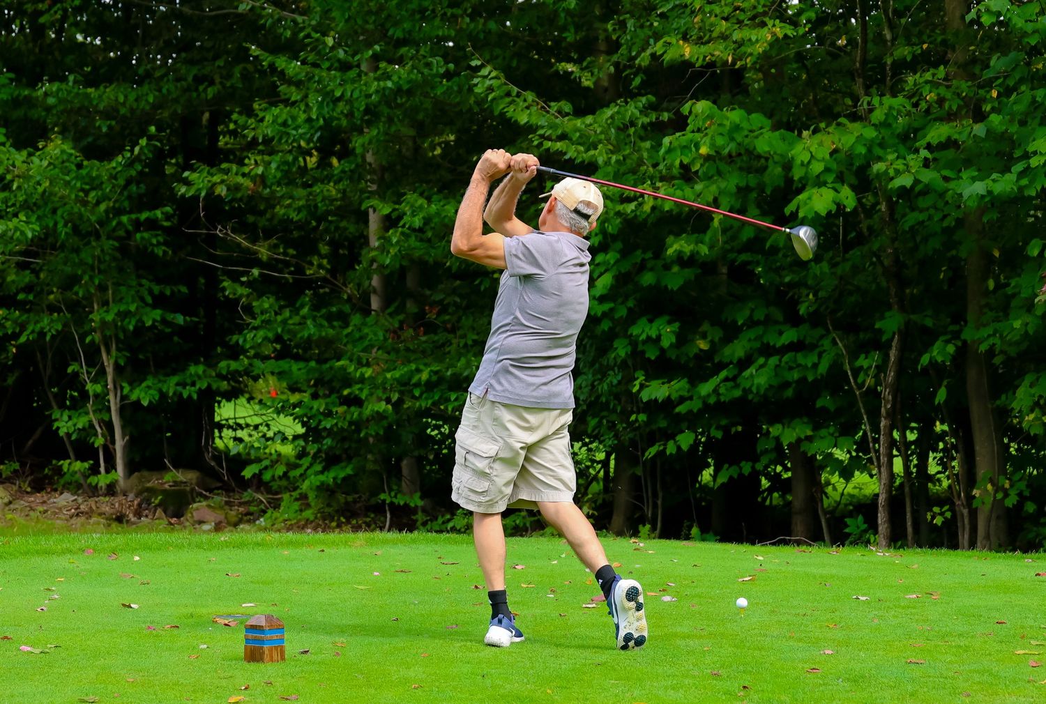 A golf ball soars through the air against a backdrop of lush green trees on a sunny day at a golf course.