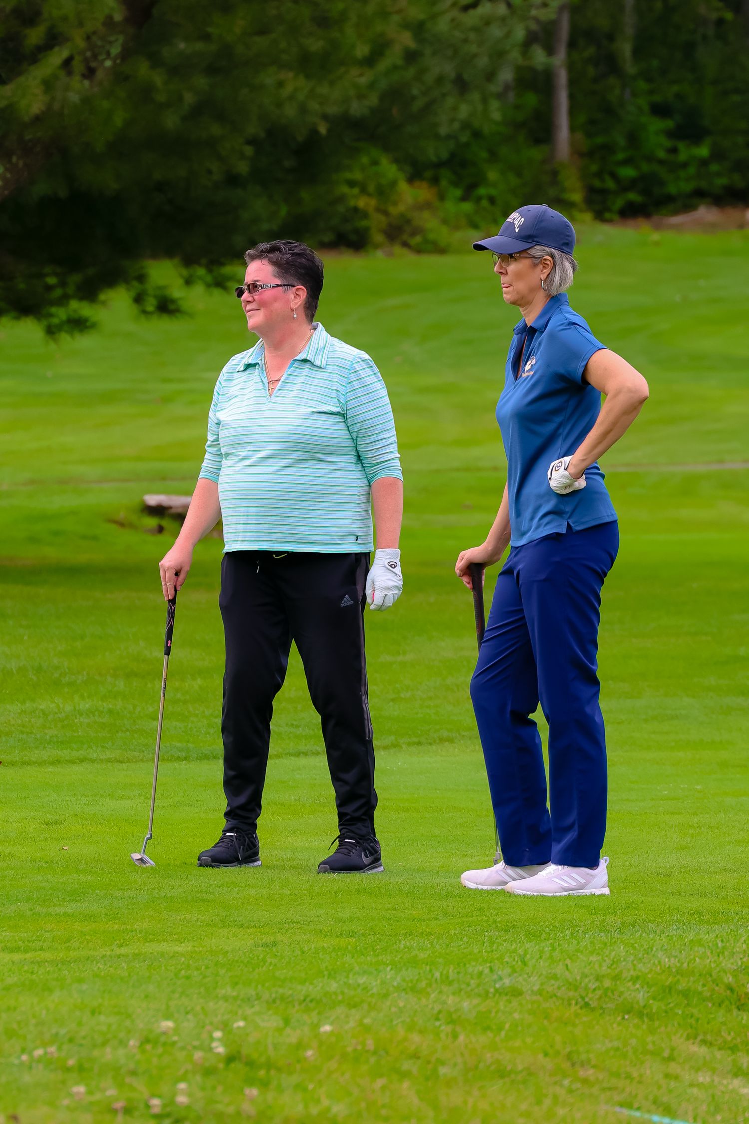 Two golfers stand on a lush green course practicing their putting technique on a sunny day.