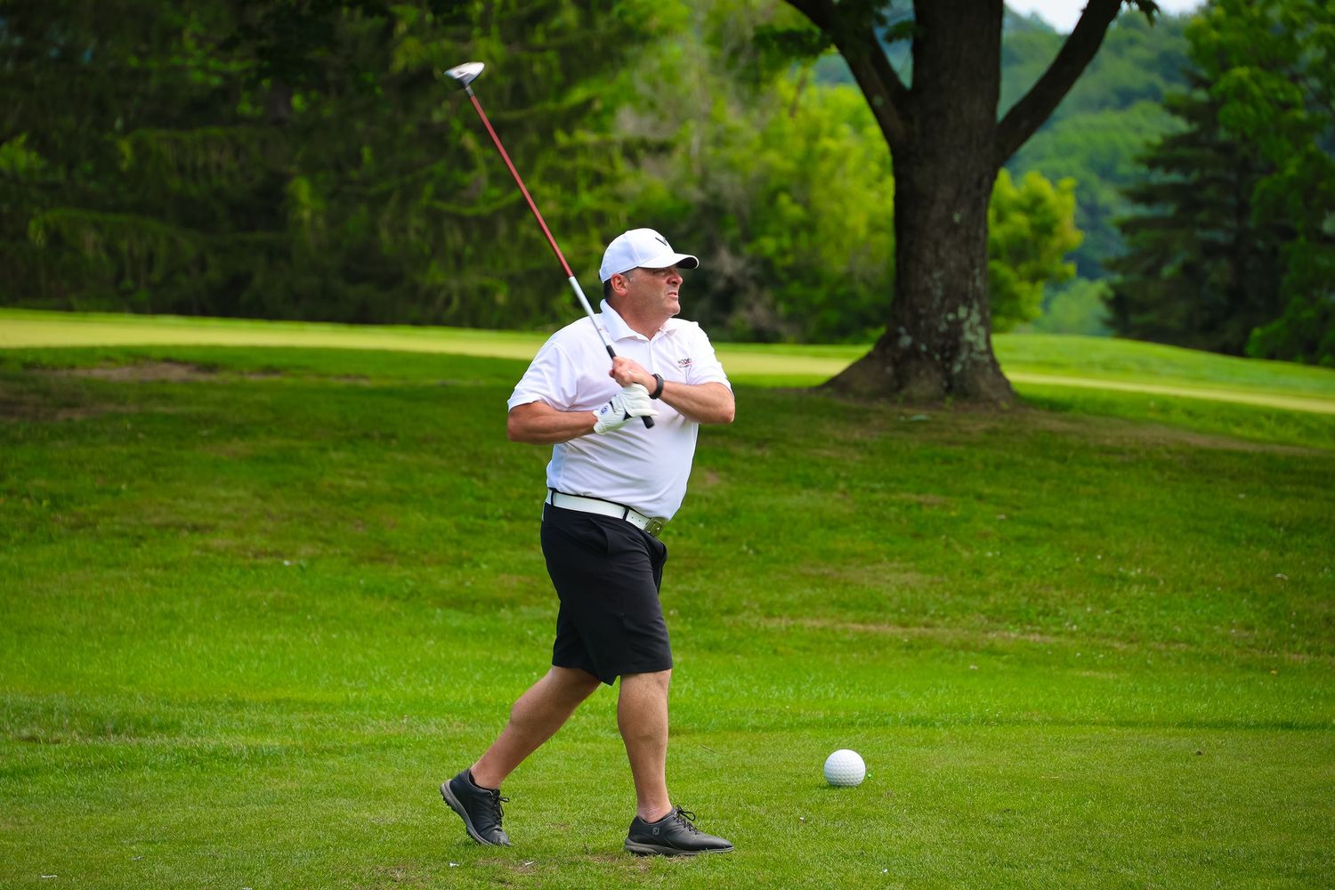 A golfer in white and black attire swings a club on a lush green golf course during a sunny day.