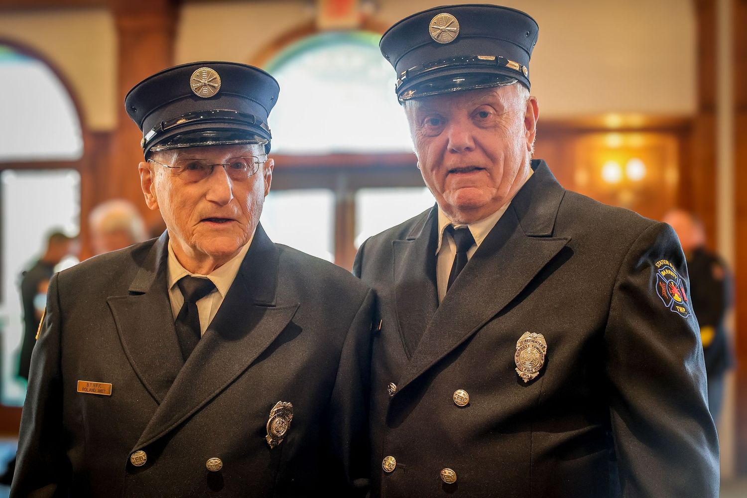 A group of uniformed fire department personnel stand together in formal dress at an indoor gathering.