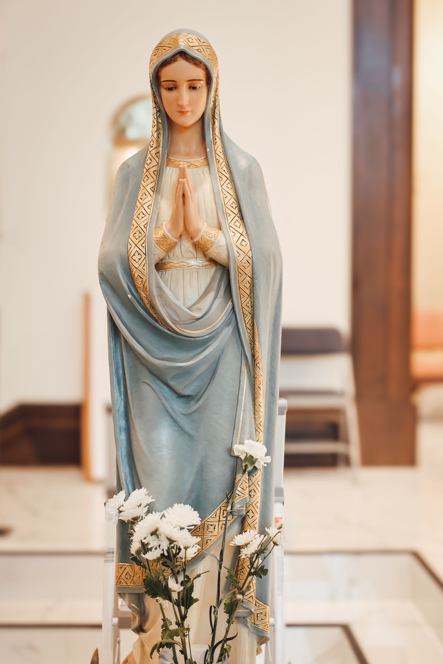 A statue of the Virgin Mary in a blue and gold robe stands gracefully with white roses at its base.