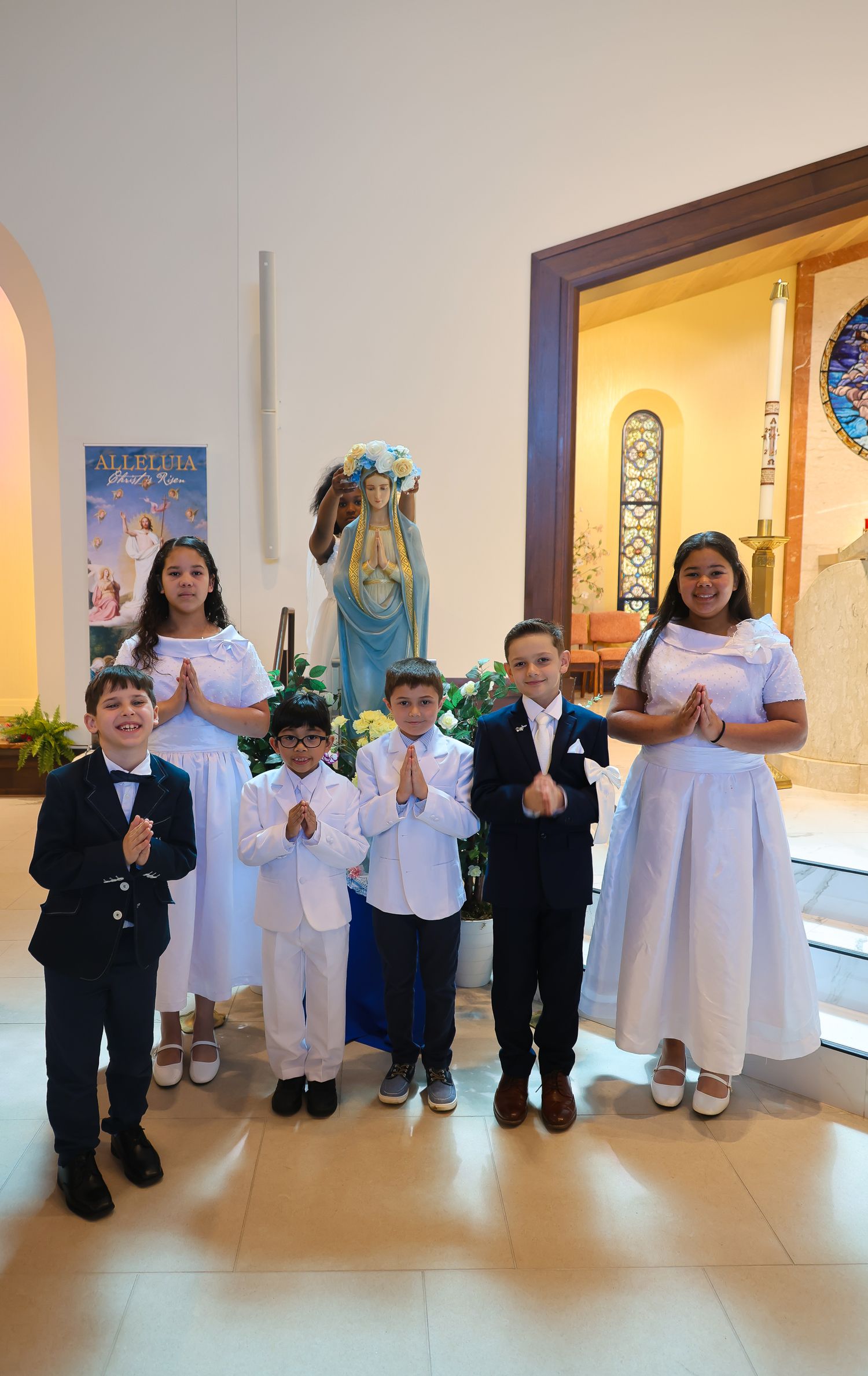 Children in white and black formal attire stand in line against a wall during a special event.
