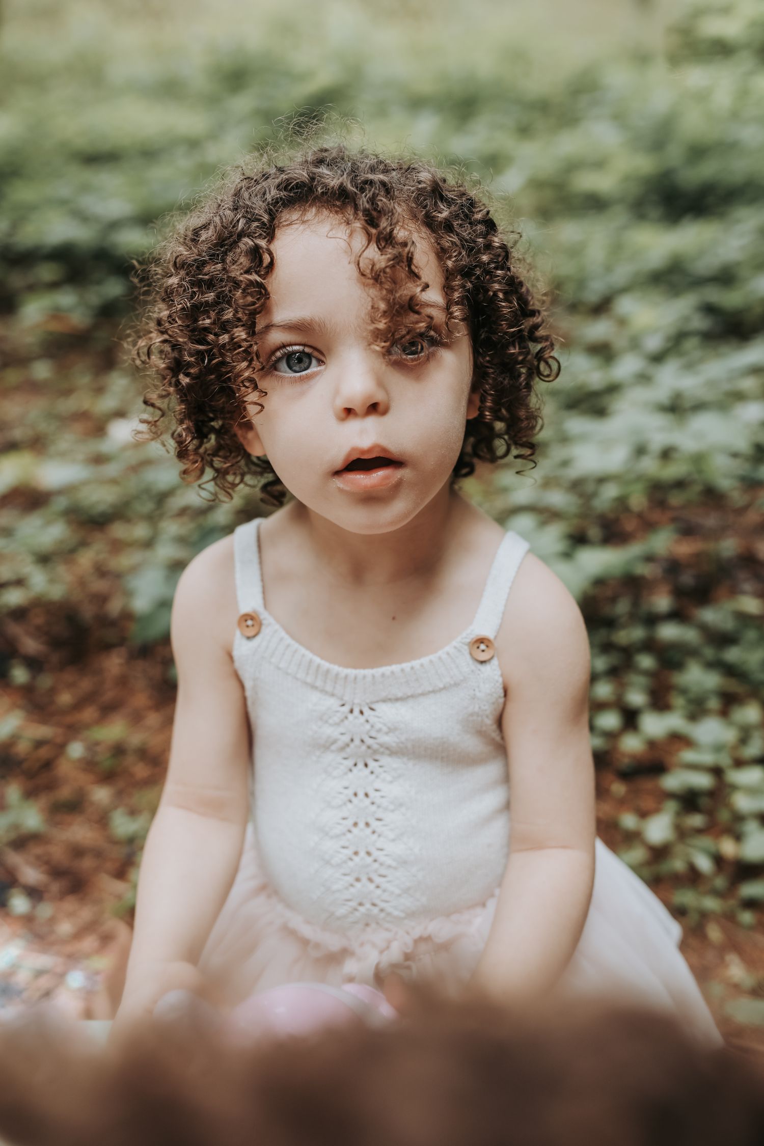 A child wearing a white lace dress sits among greenery in a dreamy outdoor setting.