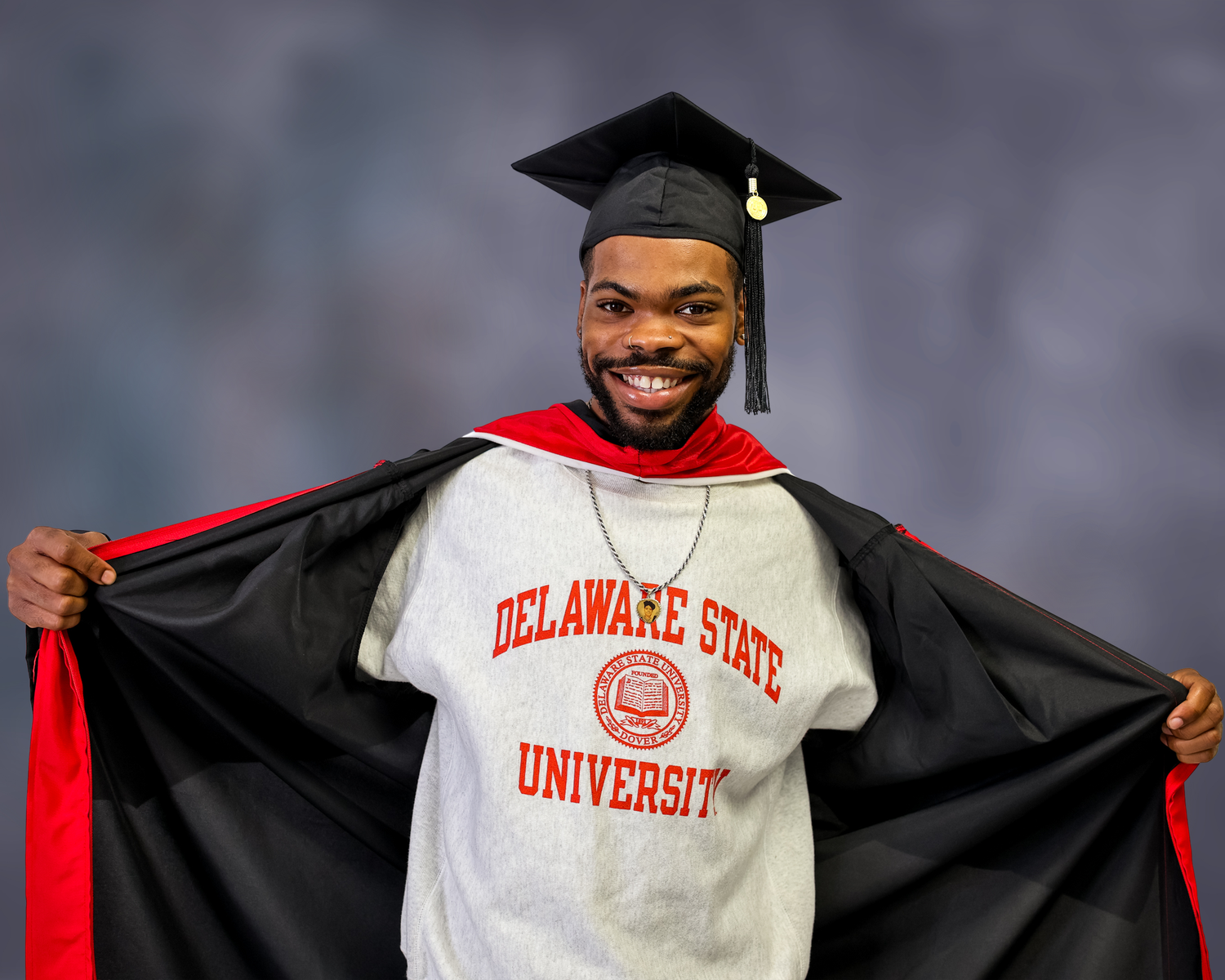Graduate in cap and gown proudly displays Delaware State University sweatshirt during graduation ceremony.
