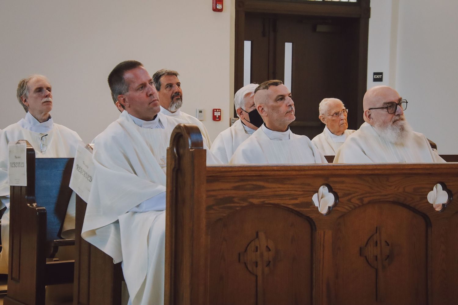 Row of clergy members in white robes seated in wooden church pews during service.