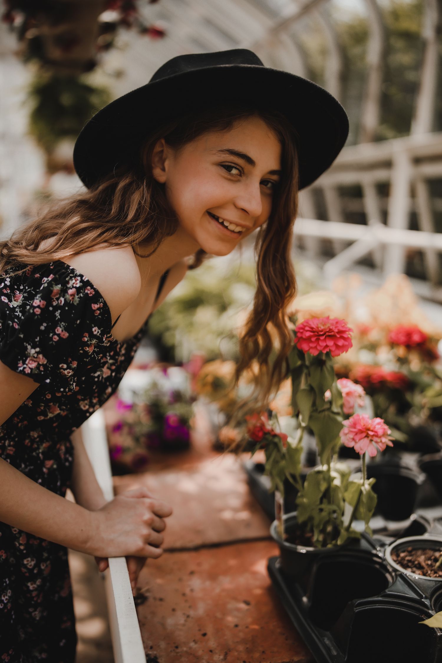 Woman in black floral dress and hat tending to colorful garden flowers.