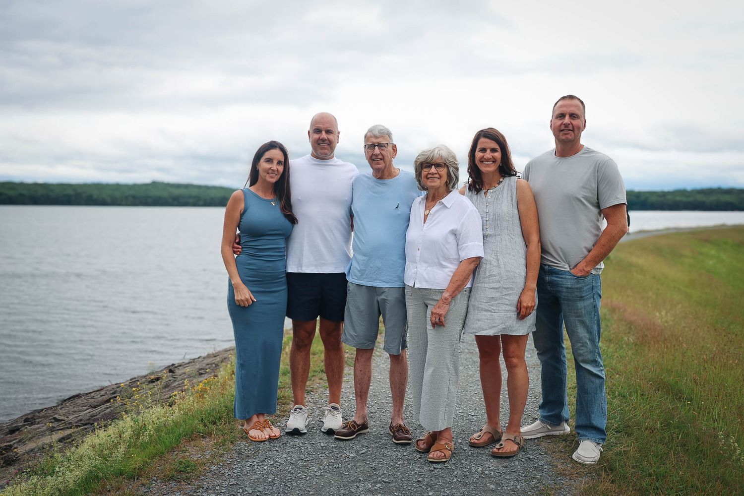 Six people pose together smiling near a scenic lakeside with green hills and cloudy skies in the background.