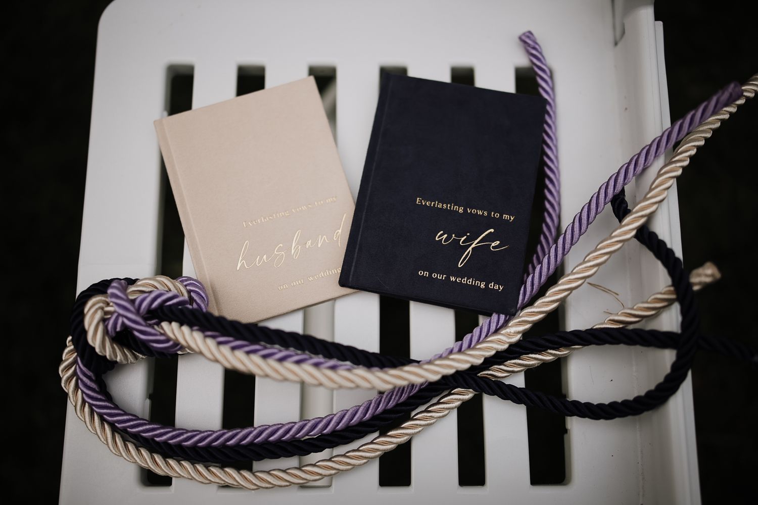 Purple and gold rope knot beside wedding vow booklets labeled husband and wife on a white chair.