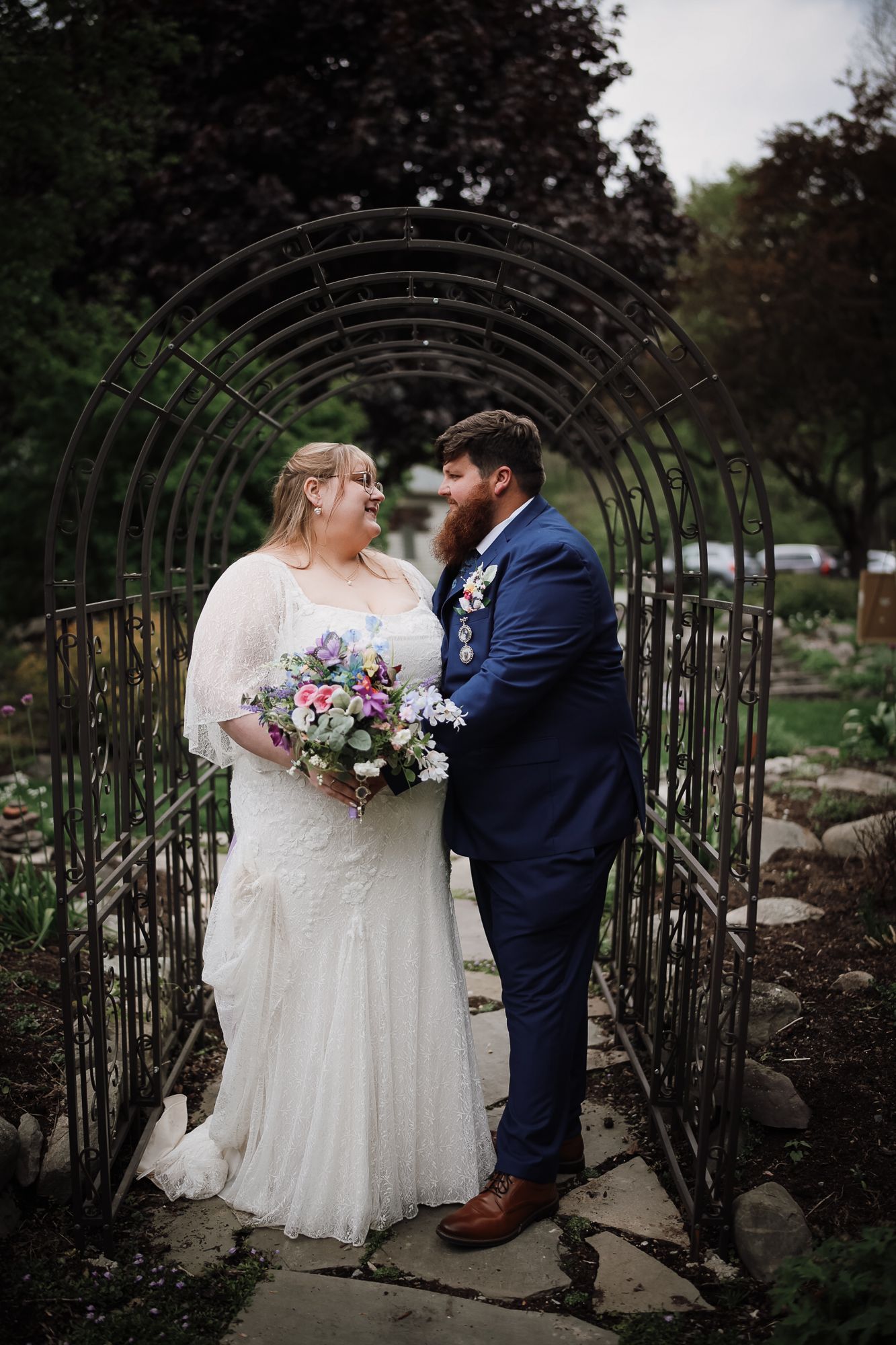 A bride and groom share a romantic moment under a metal arch in a lush garden on their wedding day.