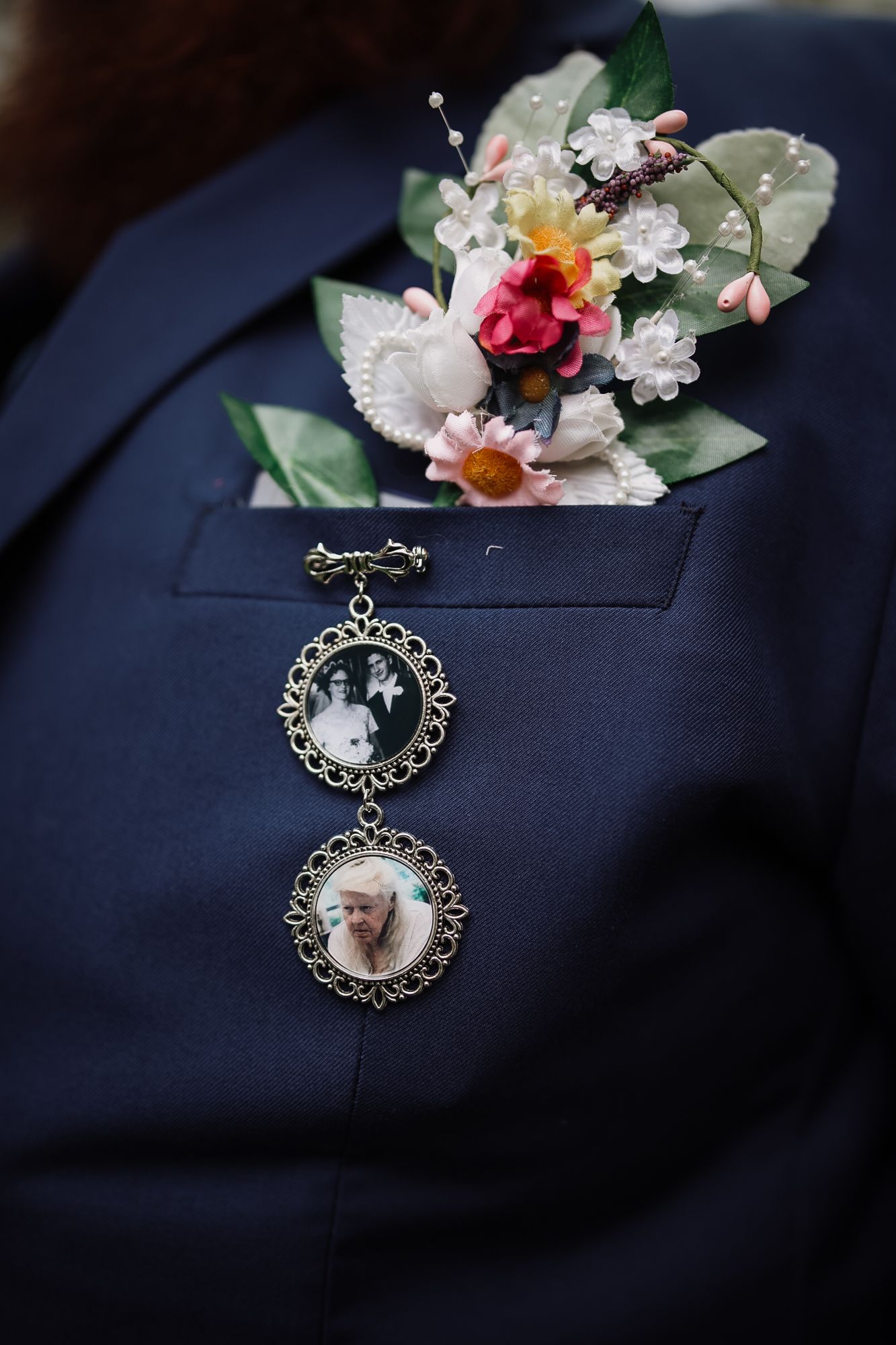 Groom's navy suit lapel with floral boutonniere and two vintage locket pins containing memorial photos of loved ones.