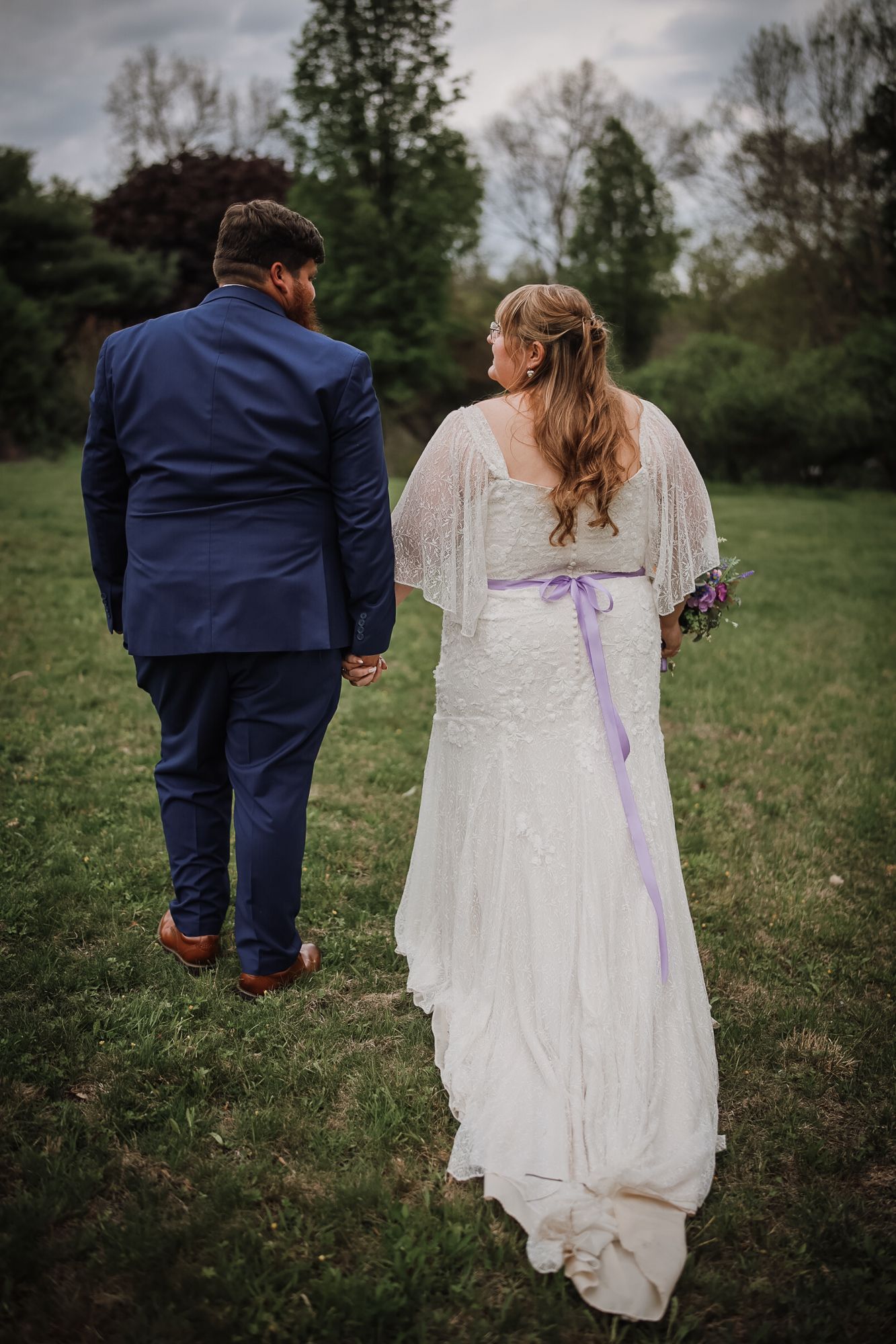 A bride and groom walk hand-in-hand through a green field, she in a white lace gown with purple sash, he in a navy suit.