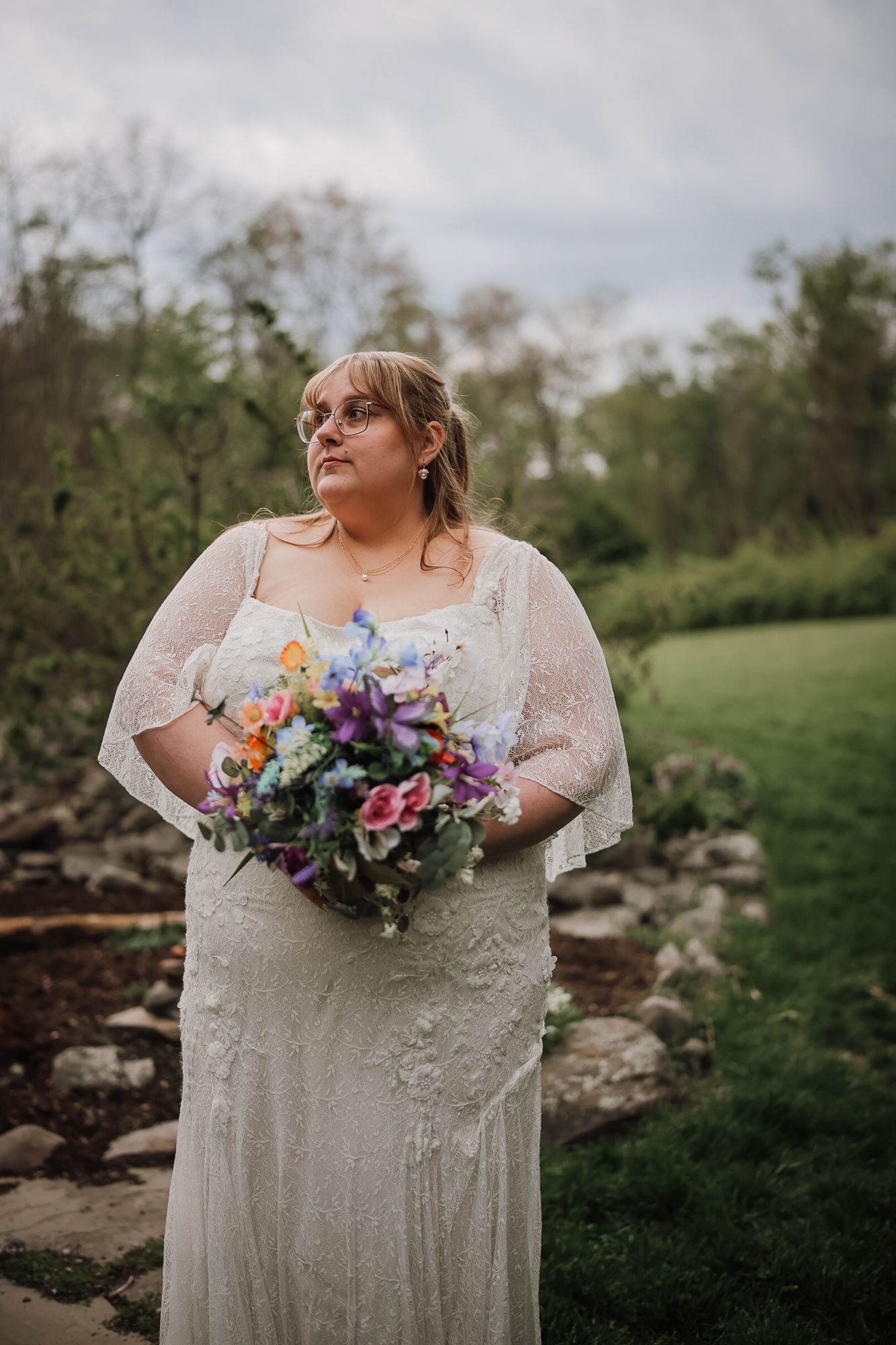 A bride in a white lace gown holds a colorful bouquet outdoors, gazing thoughtfully with a green garden backdrop.