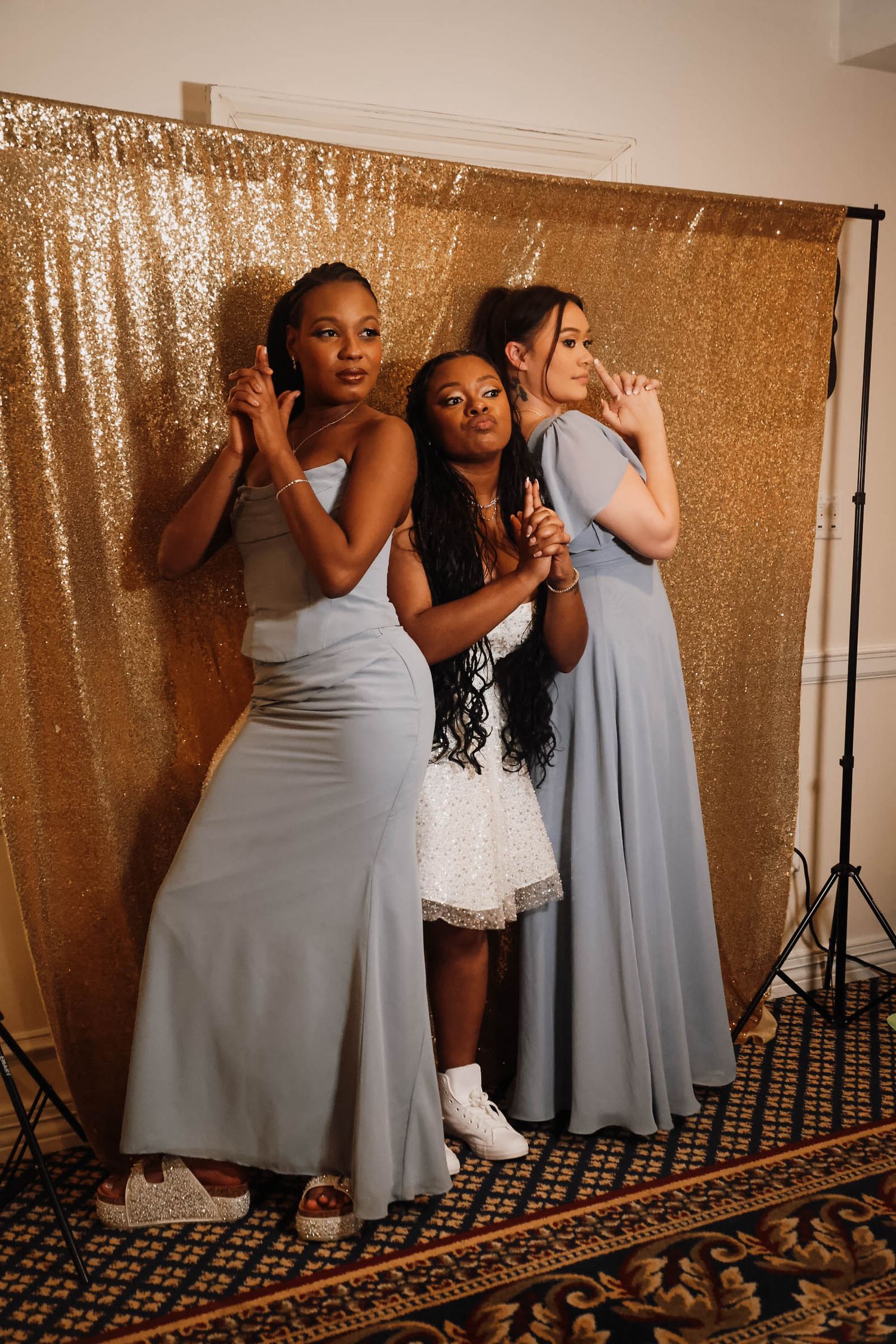 Three women in blue gowns pose elegantly before a gold sequin photo booth backdrop.