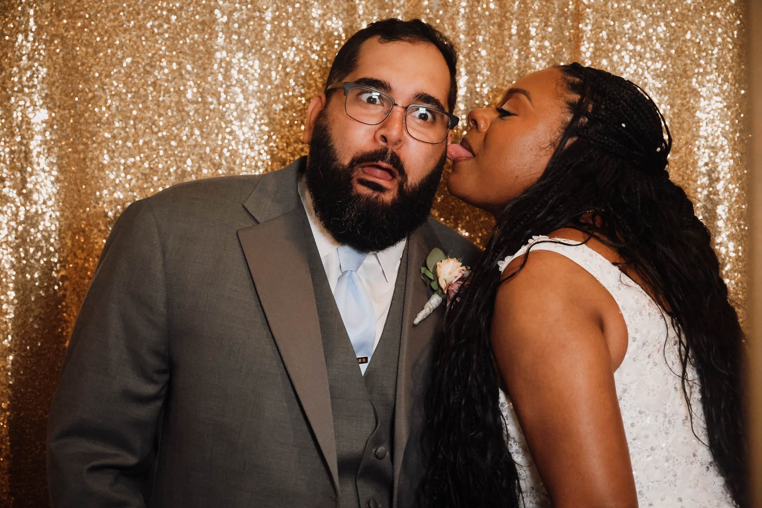 Bride in white dress kisses groom in gray suit on cheek at wedding photo booth with gold sequin backdrop.