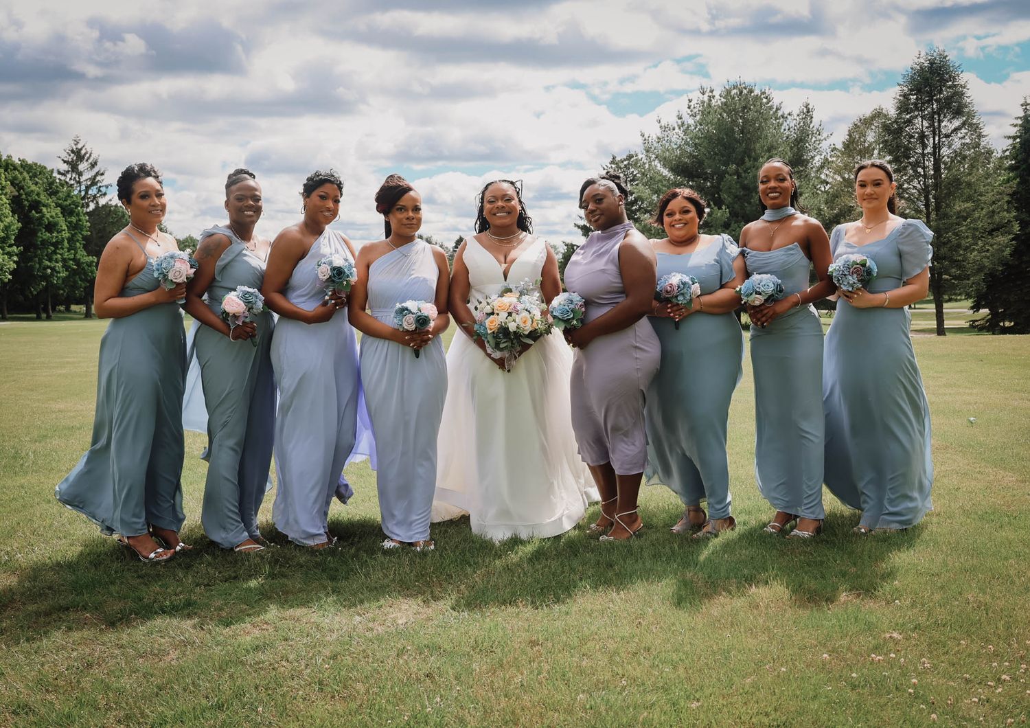Bride in white gown poses with eight bridesmaids in blue dresses holding bouquets on a green lawn.