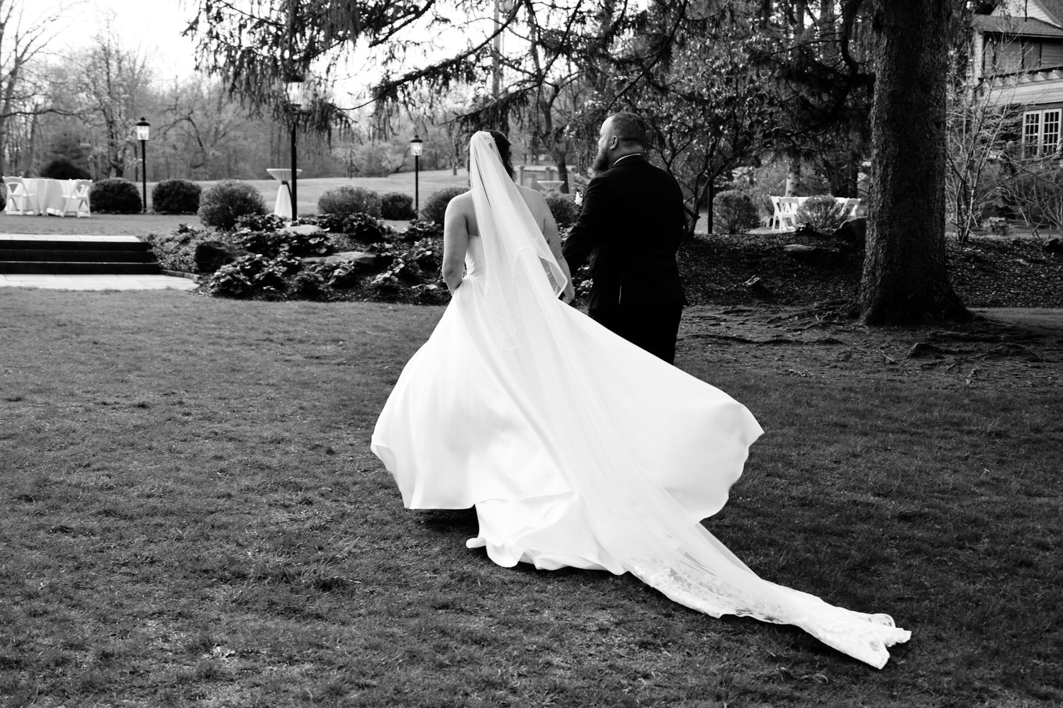 Black and white photo of a bride in a flowing white gown and veil walking with a man across a lush green lawn.
