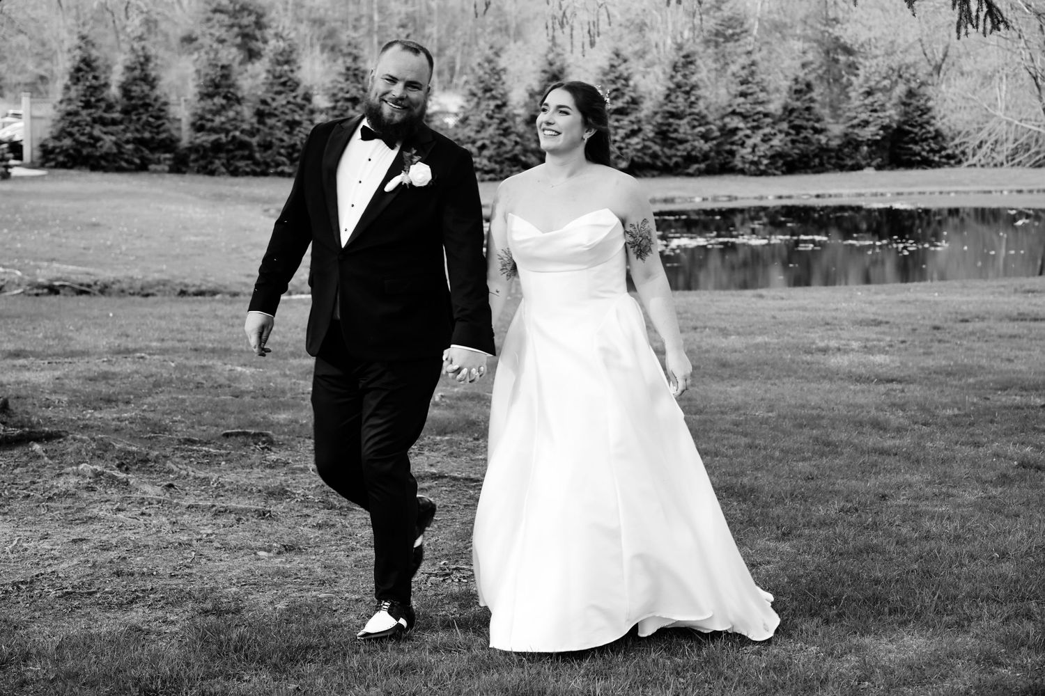 Black and white photo of a bride and groom walking hand in hand on grass near a pond on their wedding day.