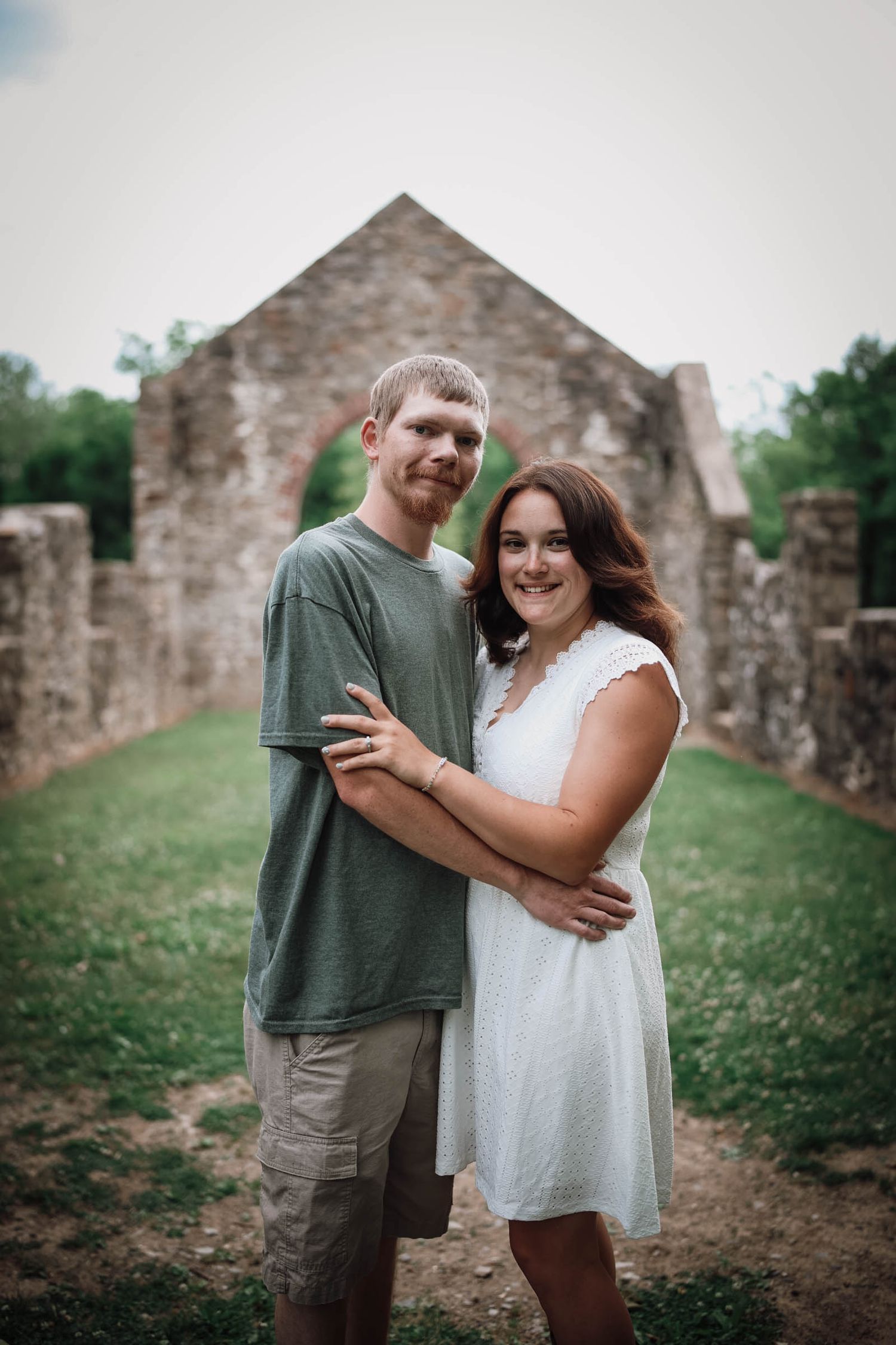 A couple embraces and smiles outdoors near rustic stone ruins, the man in a green tee and woman in a white dress.