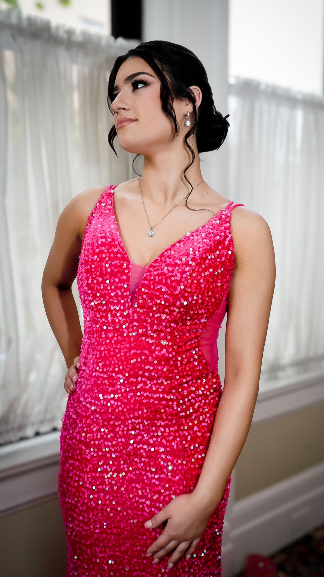 Young woman in a sparkling hot pink sequin gown with V-neckline and updo hairstyle, posing elegantly indoors.