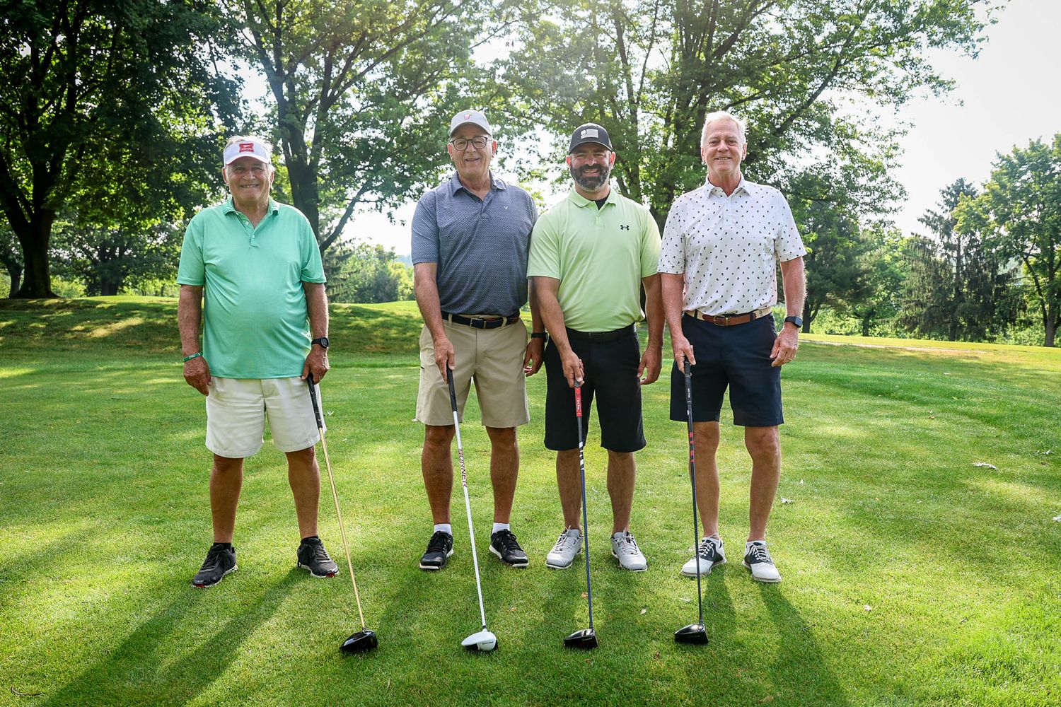 Four men posing with golf clubs on a sunny golf course surrounded by green trees.