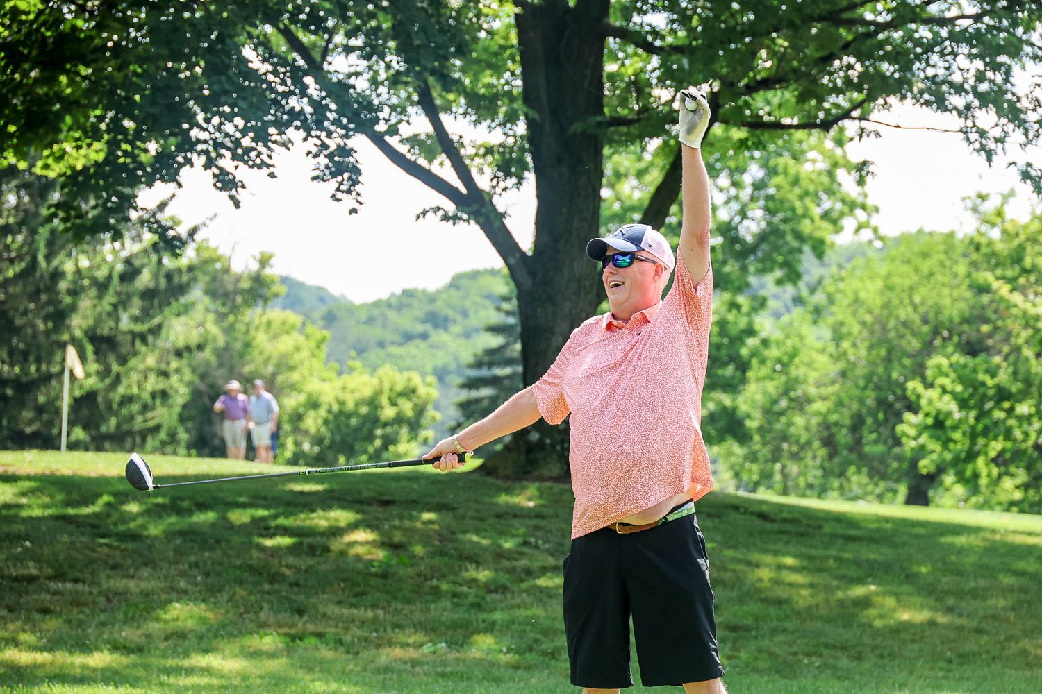 Golfer in pink shirt celebrates after a swing on a lush green golf course surrounded by trees.