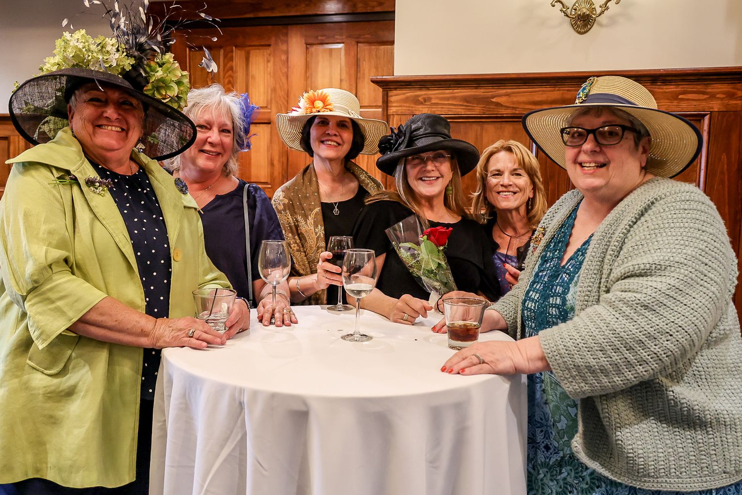 Six women wearing decorative hats pose together at a white round table, holding drinks at an elegant indoor event.